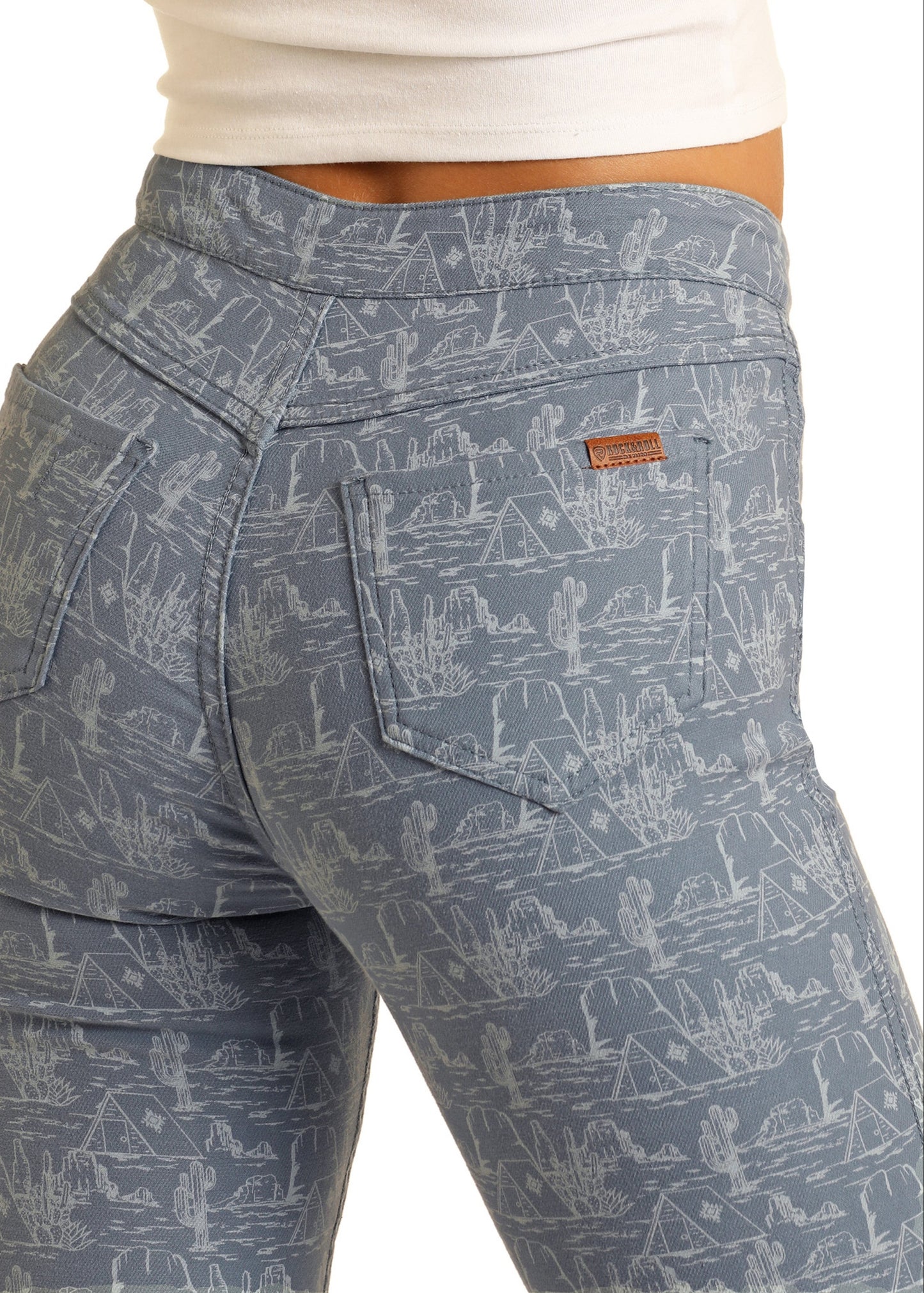 A person wears Rock & Roll Denim's Desert High Rise Reversible Flare Jeans, featuring a white desert landscape with cacti and mesas, visible back pockets, a small brown leather patch, and a white top.