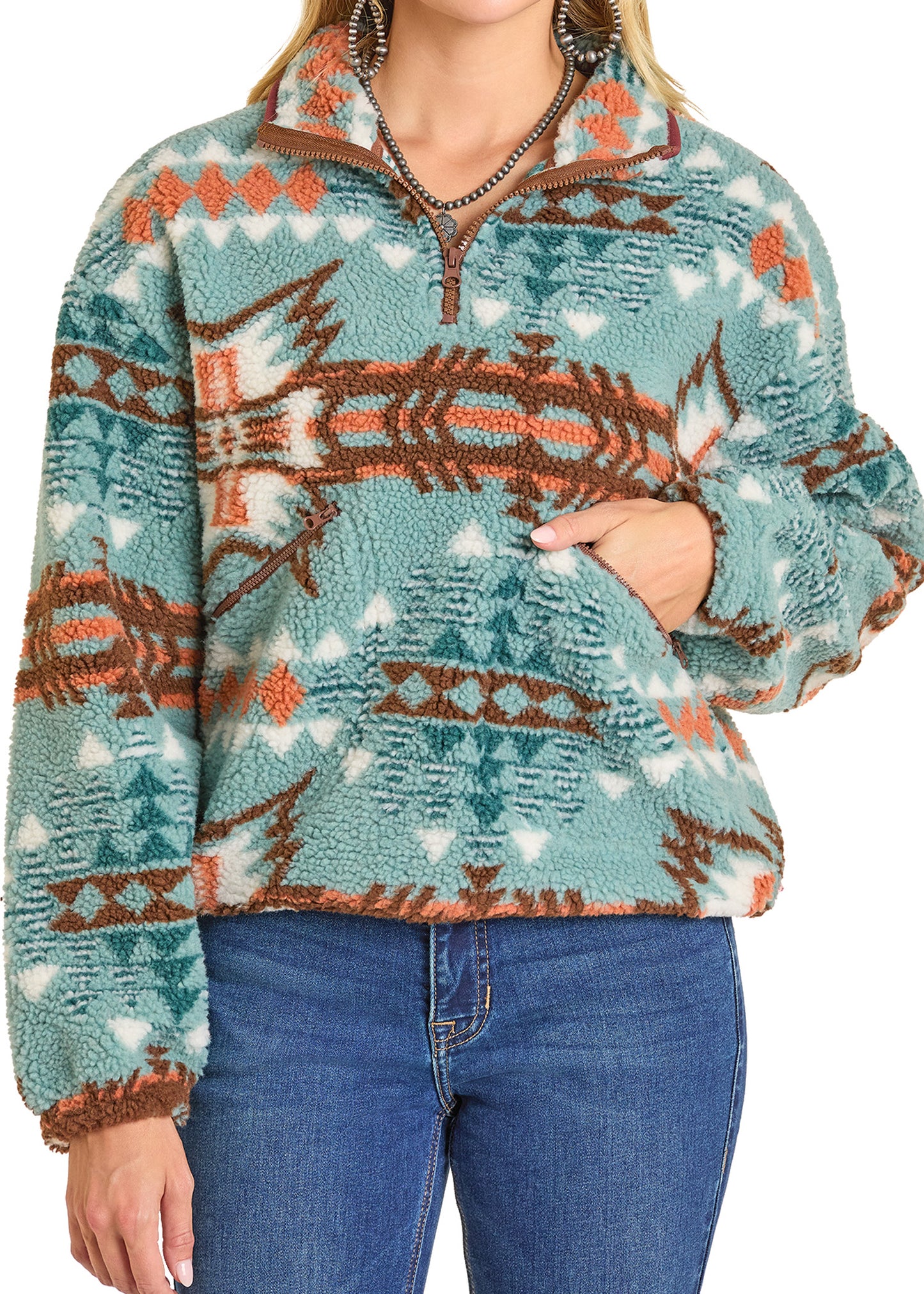 A woman wears the Rock & Roll Denim Aztec Sherpa Pullover in turquoise, brown, and white with a quarter-zip collar and blue jeans, posing with one hand in the pullover pocket.