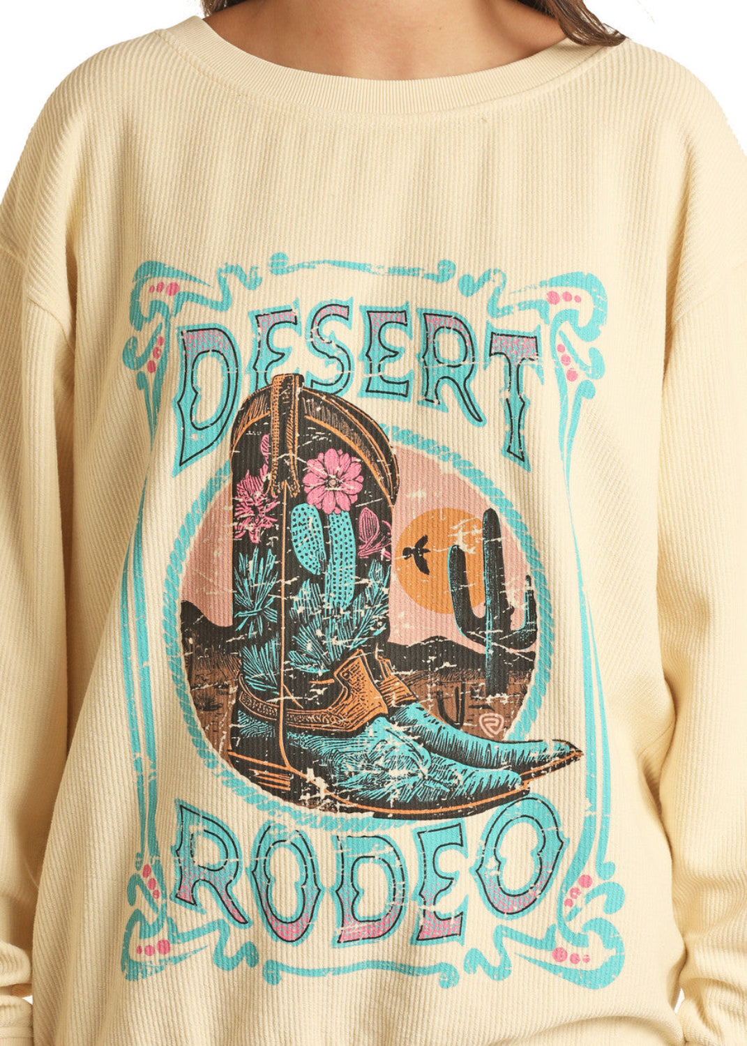 Someone wears the Rock & Roll Denim Desert Rodeo Graphic Pullover - XL, a cream sweatshirt with a bold western design of cowboy boots, cacti, desert scenery, and turquoise "DESERT RODEO" lettering for wild west flair.