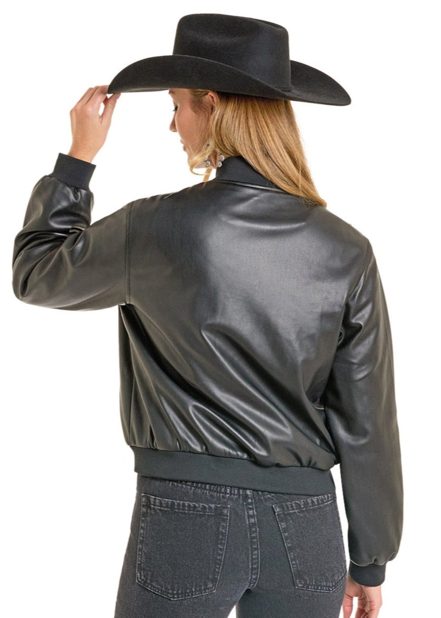 A woman in a Rock & Roll Denim Pleather Bomber Jacket, black wide-brimmed cowboy hat, and dark jeans stands with her back to the camera, glancing over her shoulder as she holds her hat brim.