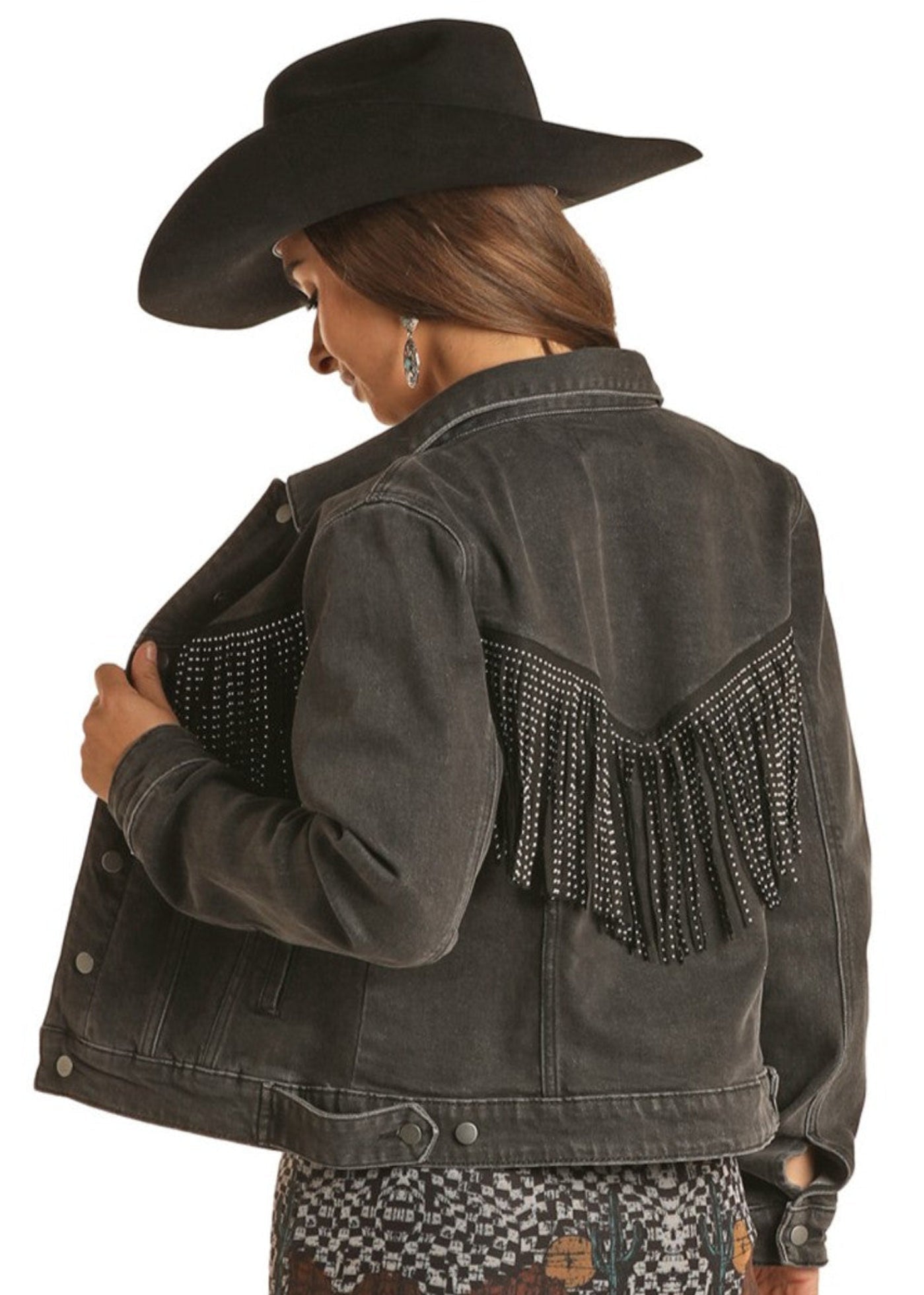 A person in a black cowboy hat and a patterned dress stands facing away, holding open one side of the Rock & Roll Denim Fringe Denim Jacket with beaded fringe details on the back.