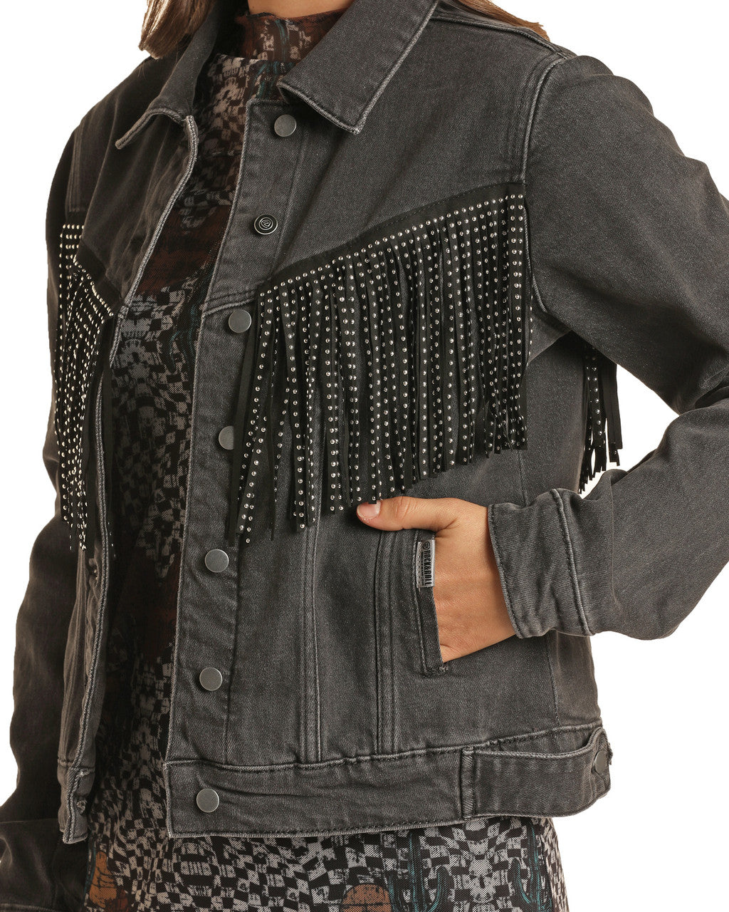 A person wearing the Rock & Roll Denim Fringe Denim Jacket, featuring studded fringe across the chest, stands with one hand in a pocket and styles the oversized jacket over a patterned dress.