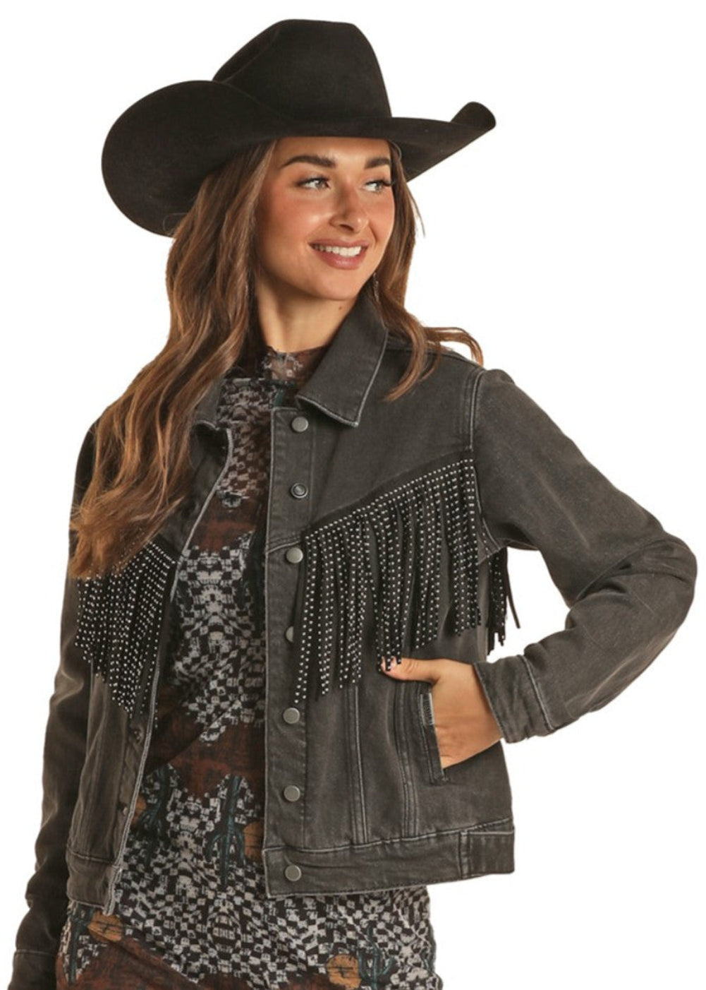 A woman in a black cowboy hat, a patterned dress, and a Rock & Roll Denim Fringe Denim Jacket with stud details stands smiling to the side with one hand on her hip.