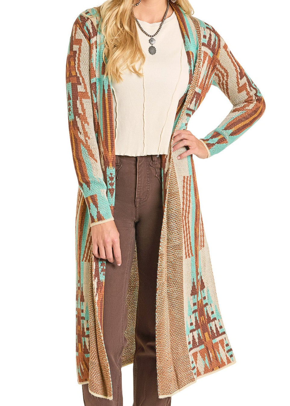 A woman with wavy blonde hair wears the Rock & Roll Denim Multicolor Aztec Knit Duster, featuring a Southwestern geometric pattern in teal, brown, and beige, over a cream top and brown pants. She accessorizes with a layered necklace.