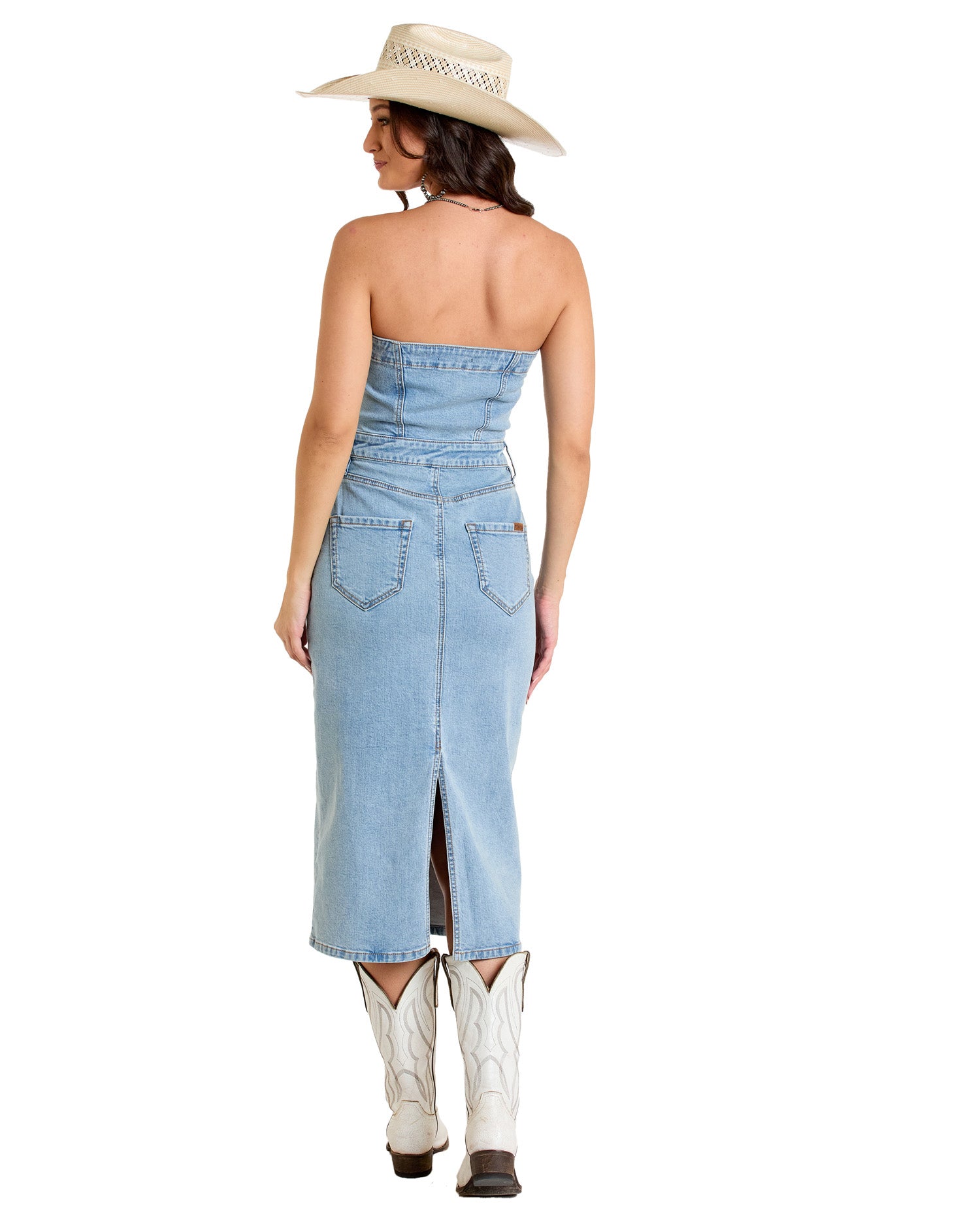 A woman faces away in the Rock & Roll Denim Detachable Strap Denim Midi Dress, paired with white cowboy boots and a tan hat. The dress features back pockets and a rear slit, showcased against a plain white background.