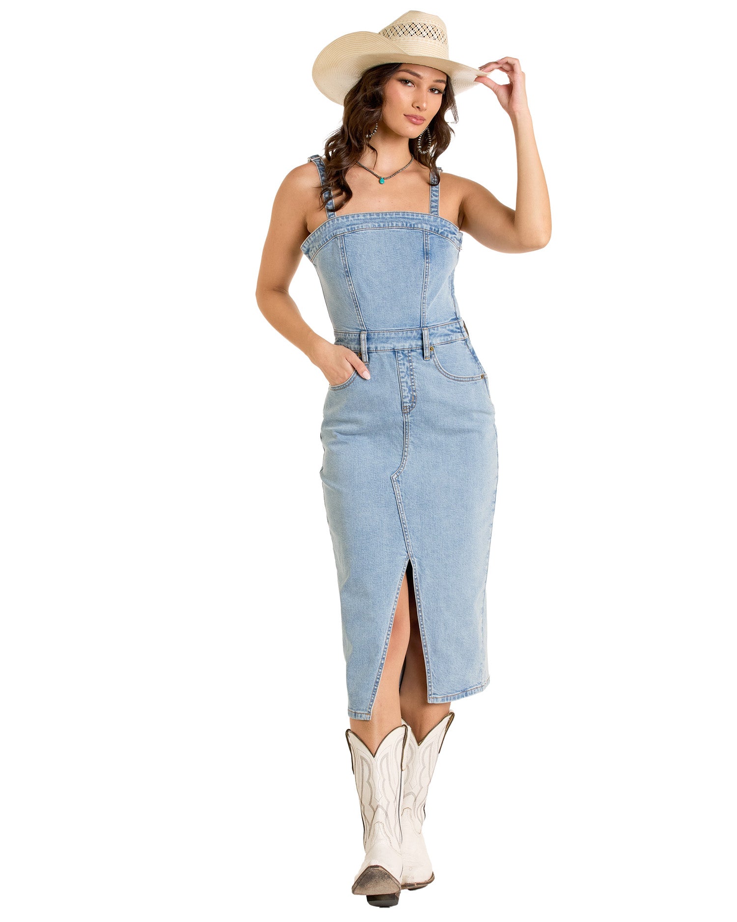 A woman smiles in the Rock & Roll Denim Detachable Strap Denim Midi Dress, styled with white cowboy boots and a hat. She stands with one hand in her pocket and the other touching her hat.