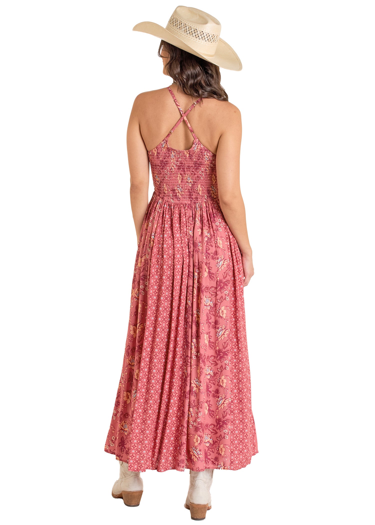 A woman in a straw hat and sleeveless Rock & Roll Denim Mixed Print Maxi Dress with crisscross straps, smocked top, and flowy skirt stands with her back to the camera, wearing white boots.