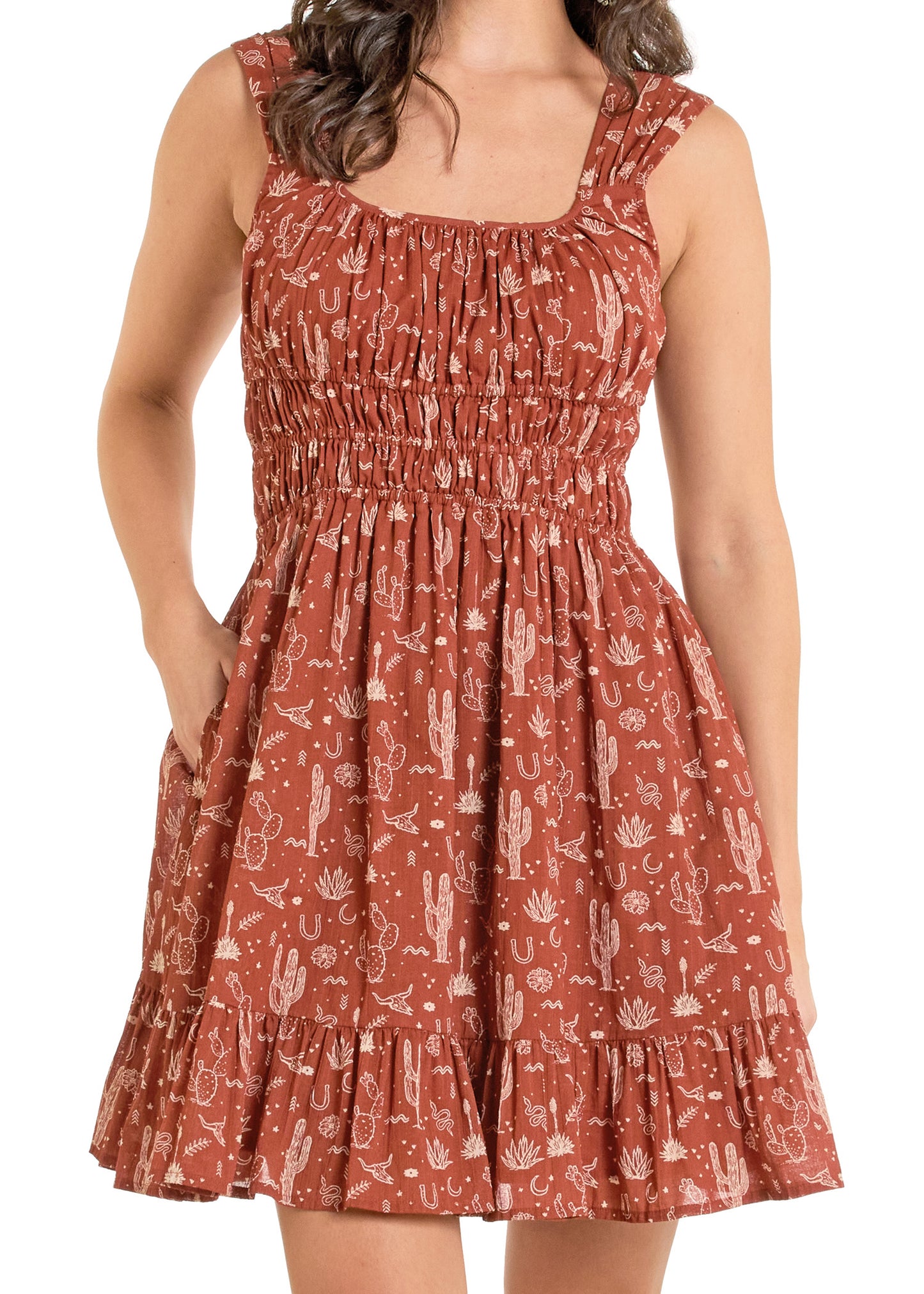 A woman wears the Rock & Roll Denim Desert Print Mini Dress, featuring a sleeveless design, rust color, white cactus and horseshoe motifs, smocked bodice, ruffled hem, and one hand in her pocket.