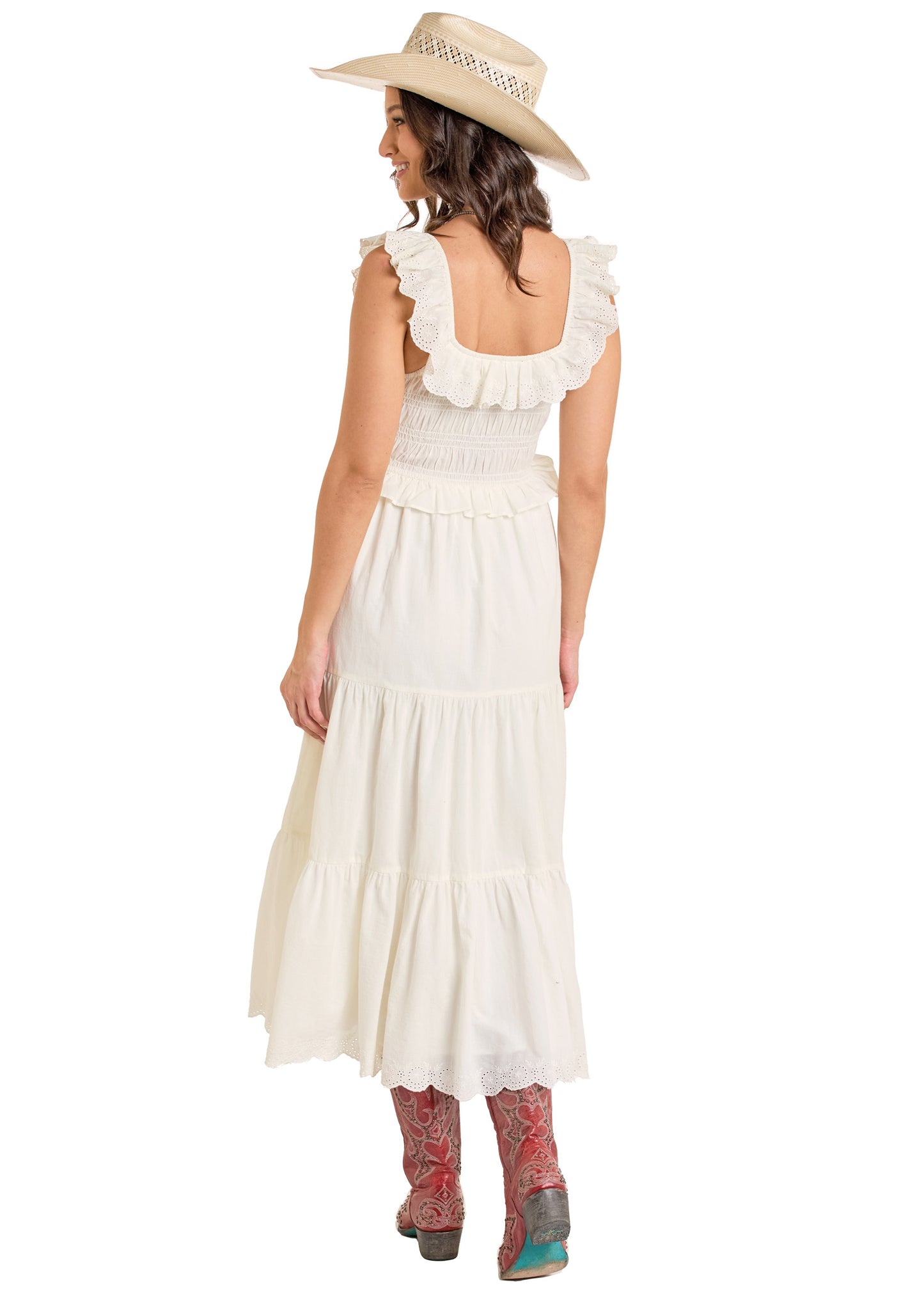 A woman wears the Rock & Roll Denim Tiered Eyelet Dress, paired with pink cowboy boots and a wide-brimmed hat. She stands against a white background, facing away and smiling over her shoulder.