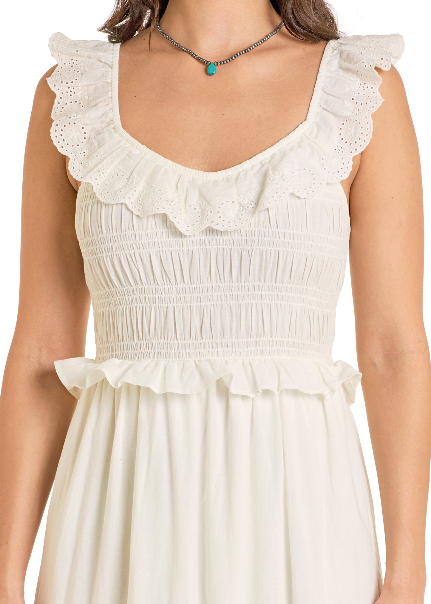 A woman in the Rock & Roll Denim Tiered Eyelet Dress, featuring white cotton fabric, ruffled lace trim at the neckline and shoulders, and a smocked top. She has long brown hair and wears a beaded necklace with a turquoise pendant.