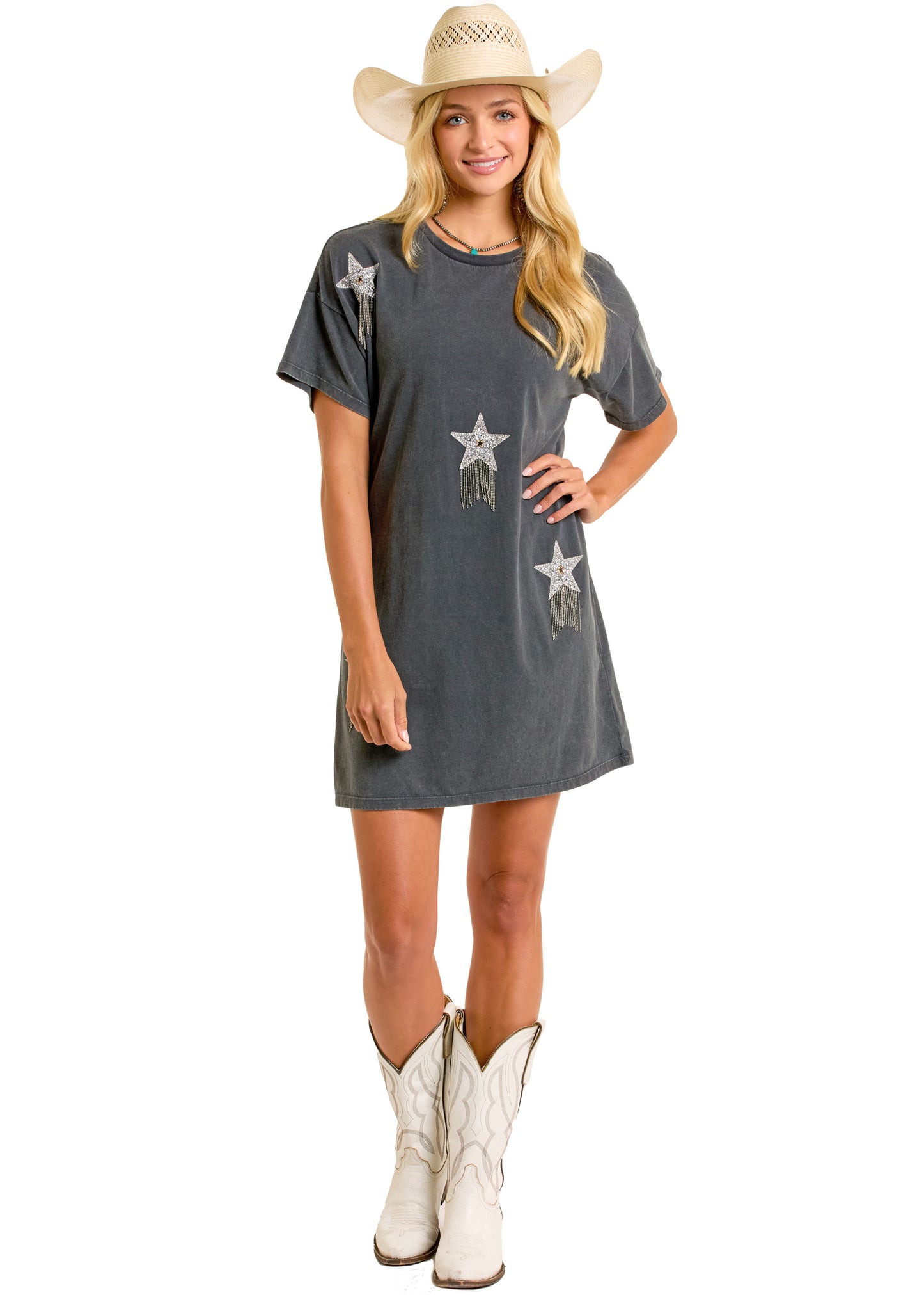 A woman in a black Rock & Roll Denim Rhinestone Star T-Shirt Dress with star, fringe, and sequin details, wearing a cowboy hat and white cowboy boots, stands smiling with her hands by her sides.