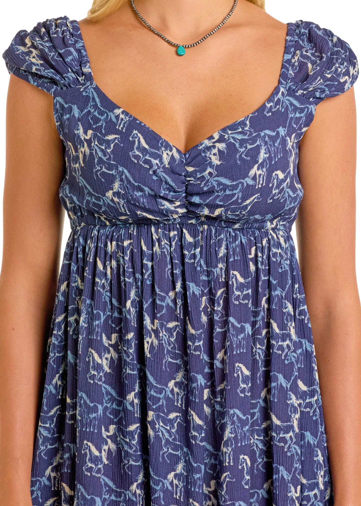 A woman wears the Rock & Roll Denim Horse Mini Dress, a blue rayon A-line with short sleeves, gathered bust, and white horse print, paired with a simple necklace featuring a turquoise bead.