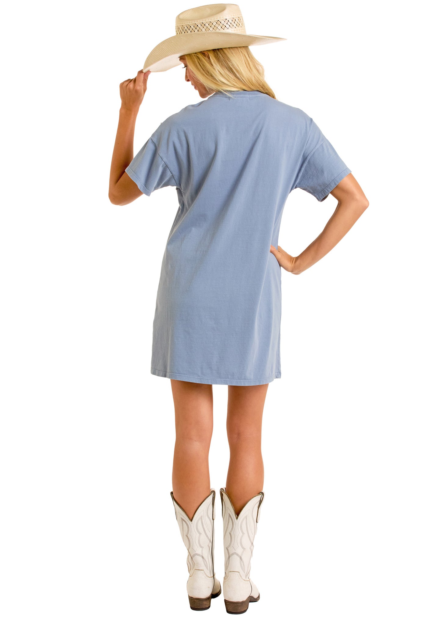 A person stands with their back to the camera, hand on hip and looking slightly to the side, wearing the Rock & Roll Denim Rhinestone T-Shirt Dress in light blue, white cowboy boots, and a straw cowboy hat.