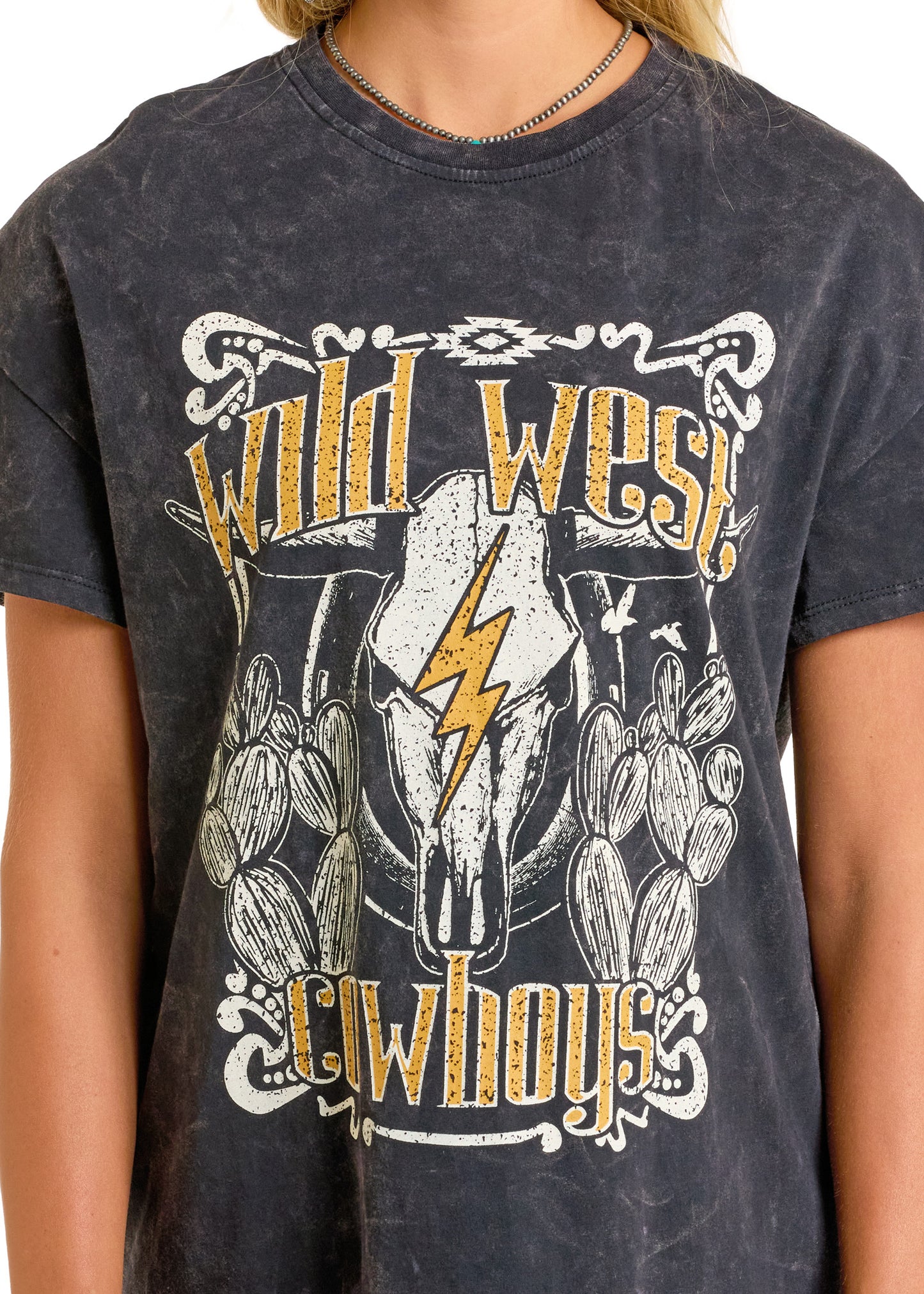 A person wearing the Rock & Roll Denim Wild West Cowboys T-Shirt Dress, a black western-style dress featuring a cow skull with a yellow lightning bolt, cacti, and bold "Wild West Cowboys" graphics.
