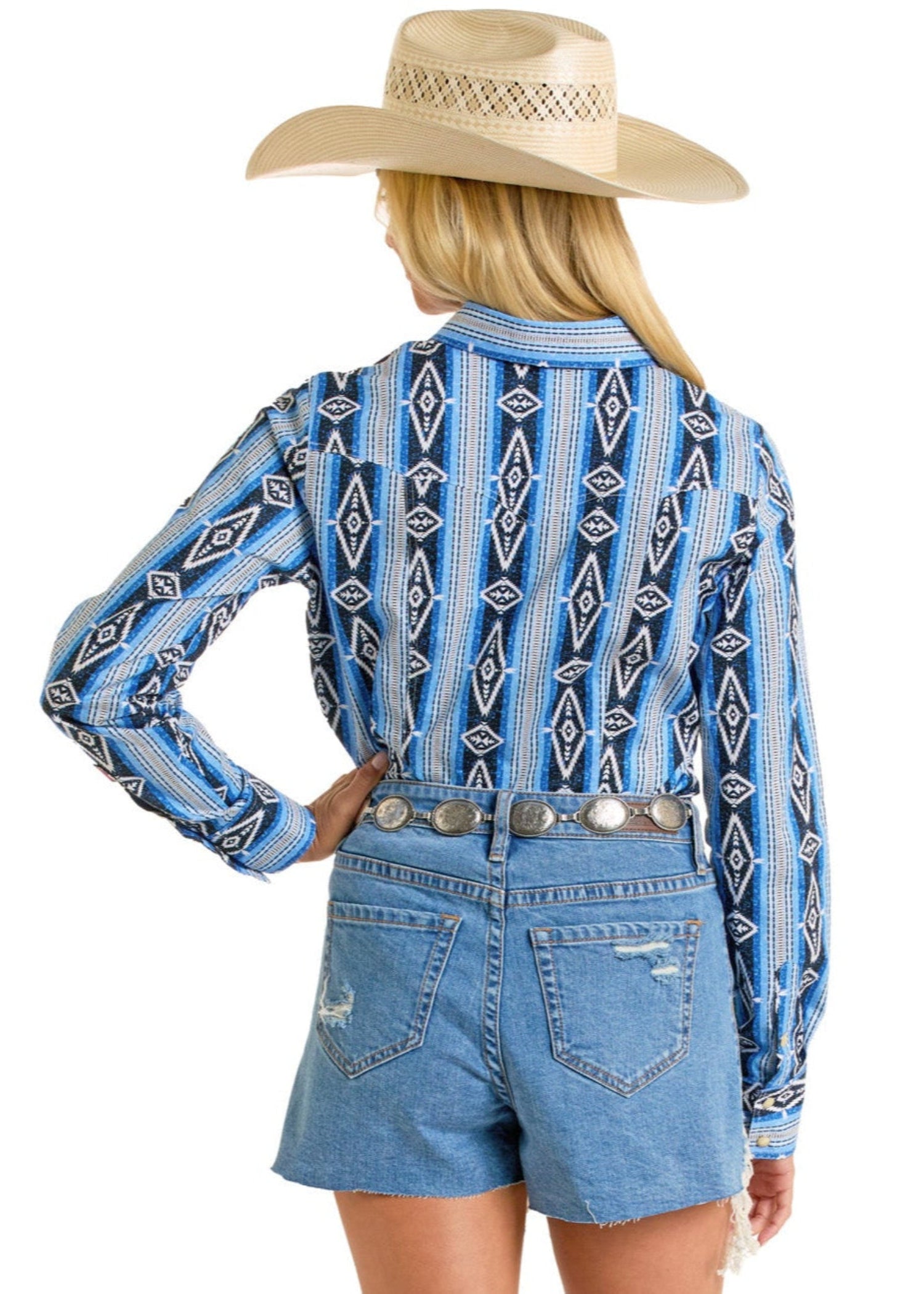 A woman wearing the Rock & Roll Denim Aztec Print Long Sleeve Snap Shirt, a wide-brimmed straw cowboy hat, and high-waisted denim shorts with a decorative belt stands facing away against a white background.