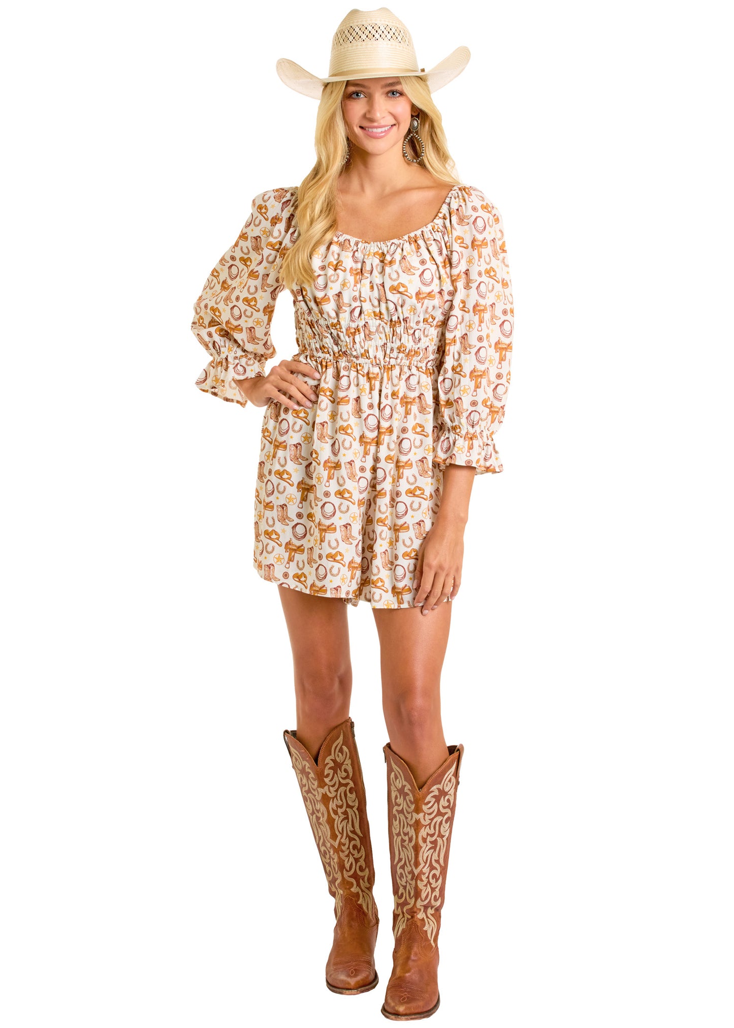 A smiling woman stands facing forward with one hand on her hip, wearing the Rock & Roll Denim Conversational Print Romper, a white cowboy hat, and tall brown cowboy boots.