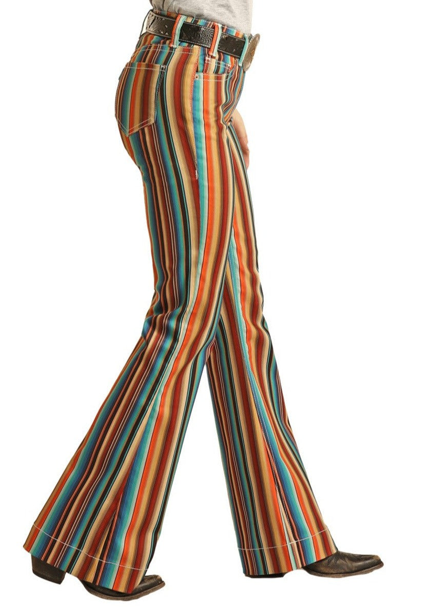 A person walks left wearing Rock & Roll Denim's Hooey Striped High Rise Trouser Jeans in vibrant orange, blue, and yellow tones with brown shoes. Only their lower body is visible, showing off a colorful western-inspired look.
