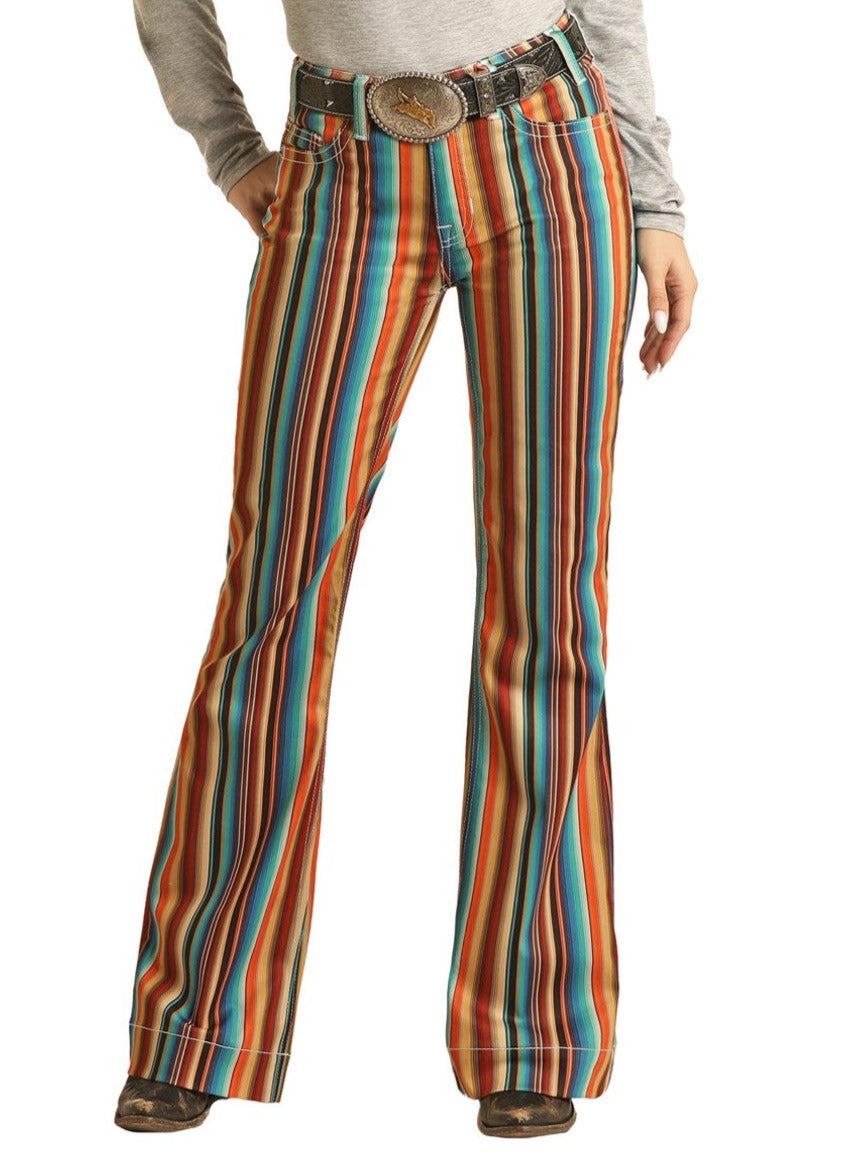 Exuding western charm, this person wears Rock & Roll Denim's Hooey Striped High Rise Trouser Jeans, a grey long-sleeve shirt, a large western belt buckle, and brown shoes, confidently posing with one hand in their pocket.