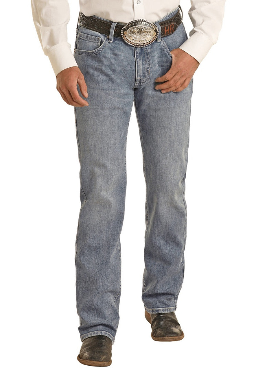 A man wearing light blue Rock & Roll Denim Vintage '46 Regular Fit V Embroidered Stackable Bootcut Jeans, a white shirt, a large belt with an ornate buckle, and black cowboy boots stands against a plain background.