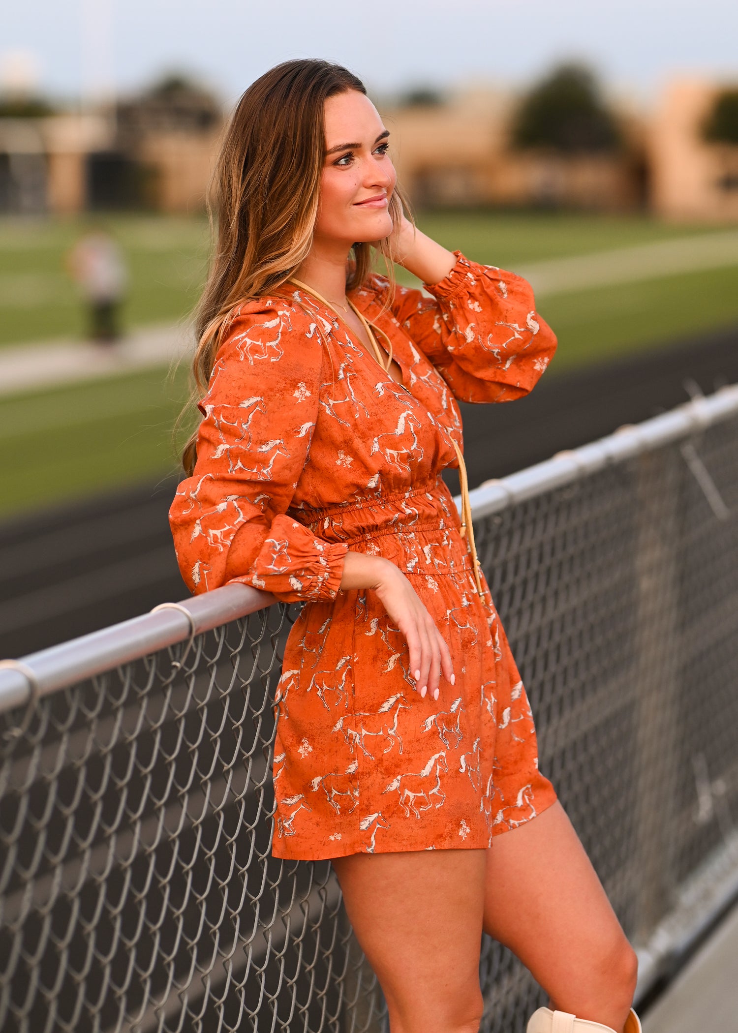 A young woman in a Horse Print Romper by Rock & Roll Denim leans on a chain-link fence by a track, smiling and looking away, with blurred buildings and greenery in the background.