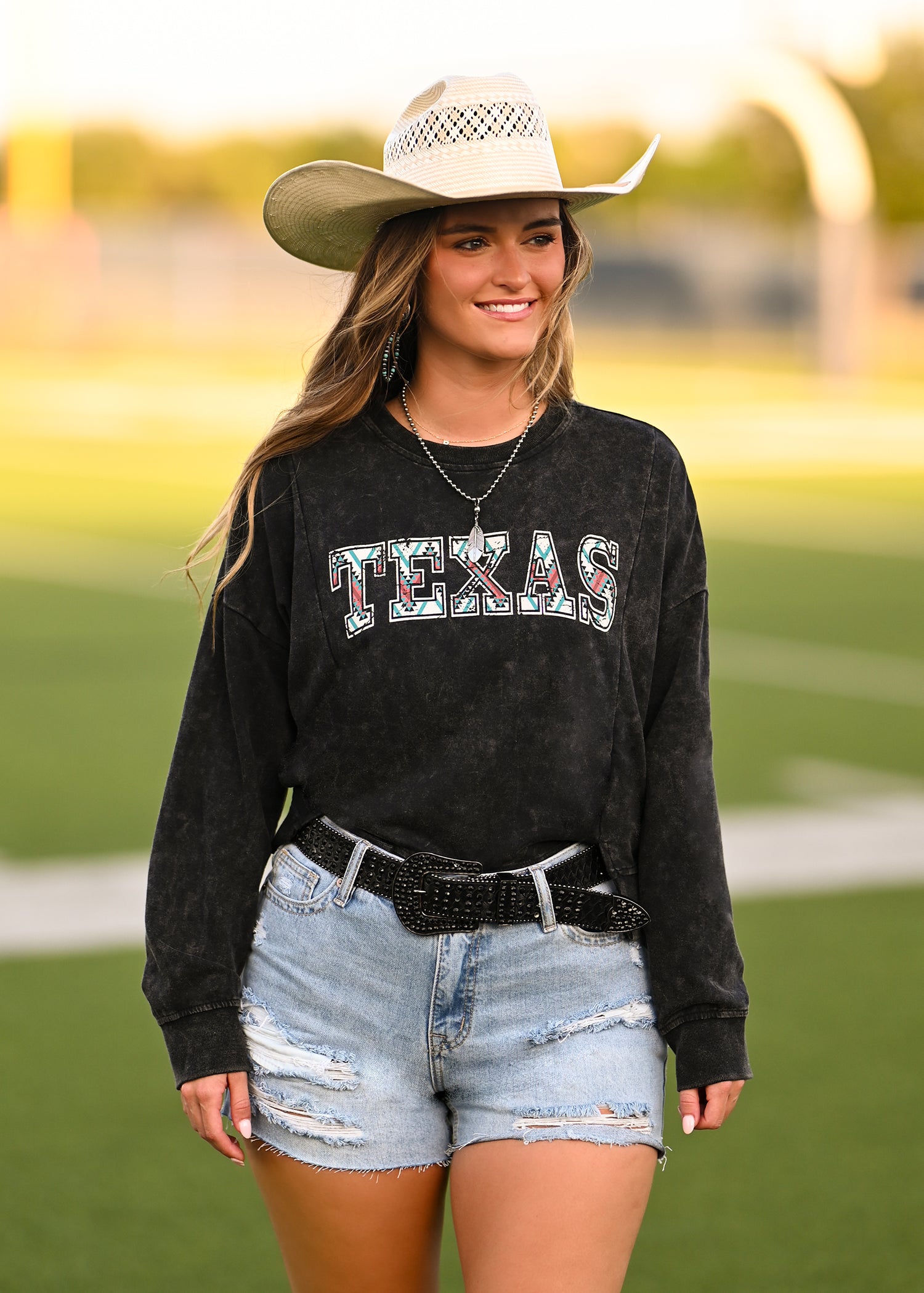 A woman in a white cowboy hat, denim shorts, and a black belt stands smiling on a football field, wearing the Rock & Roll Denim Texas Graphic Pullover.