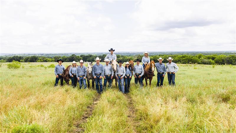 Group of people in cowboy hats and jeans standing in a grassy field with horses.