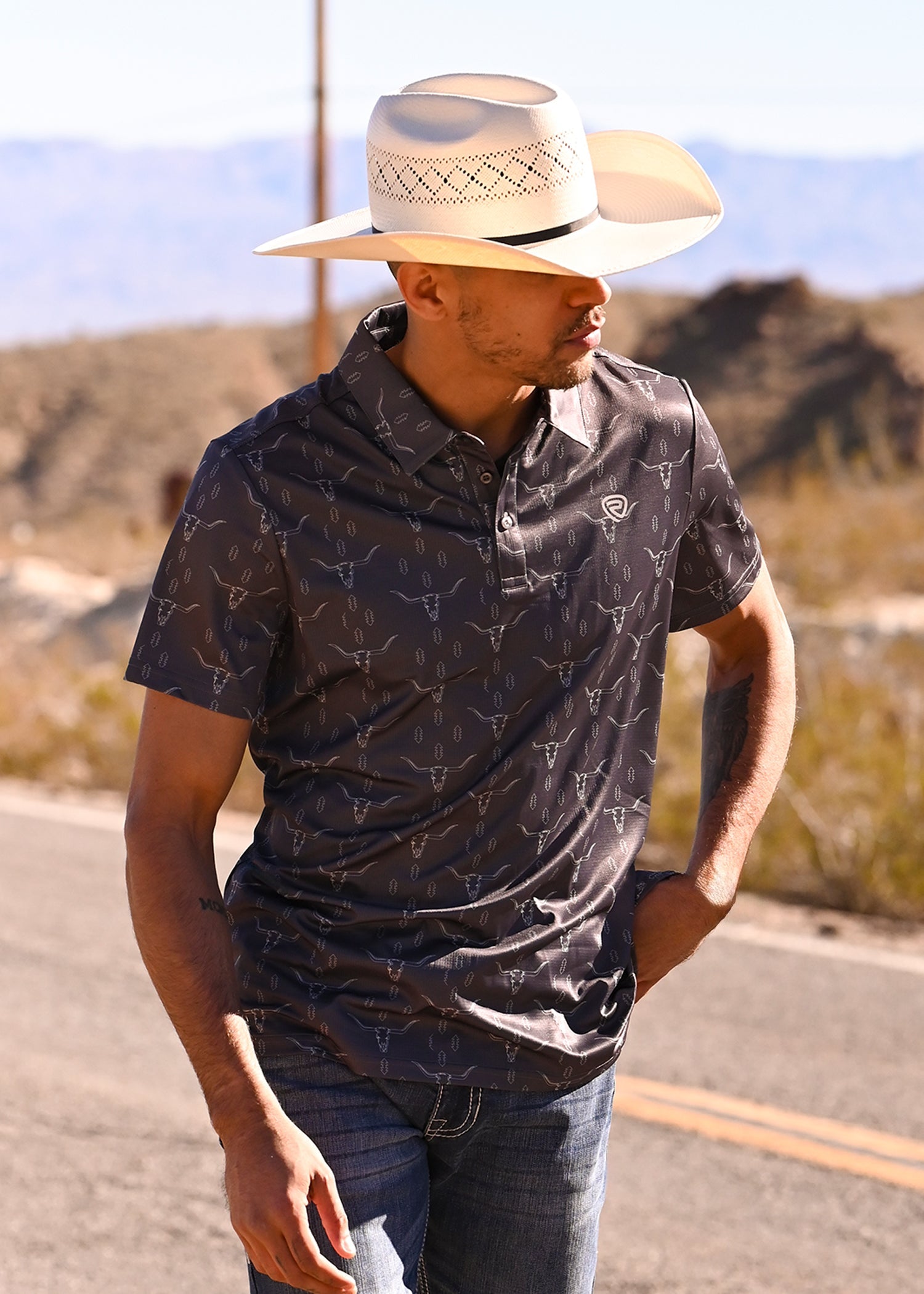 A man wearing a beige cowboy hat, a navy blue patterned polo shirt, and jeans stands smiling, looking to his left against a plain light background.
