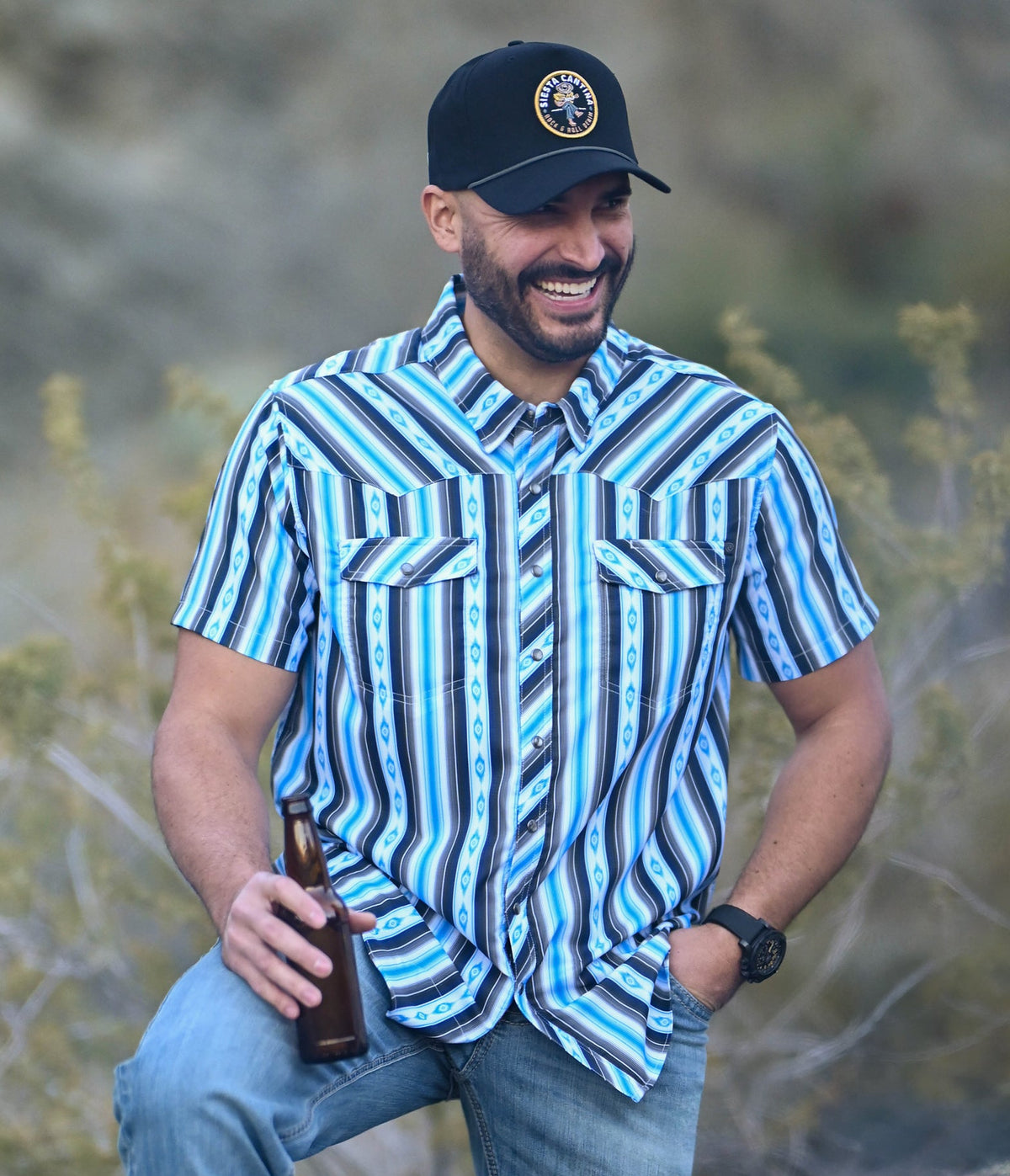 A smiling man wearing a light tan cowboy hat and a turquoise short-sleeve button-up shirt with a geometric pattern stands with his left hand in his jeans pocket.
