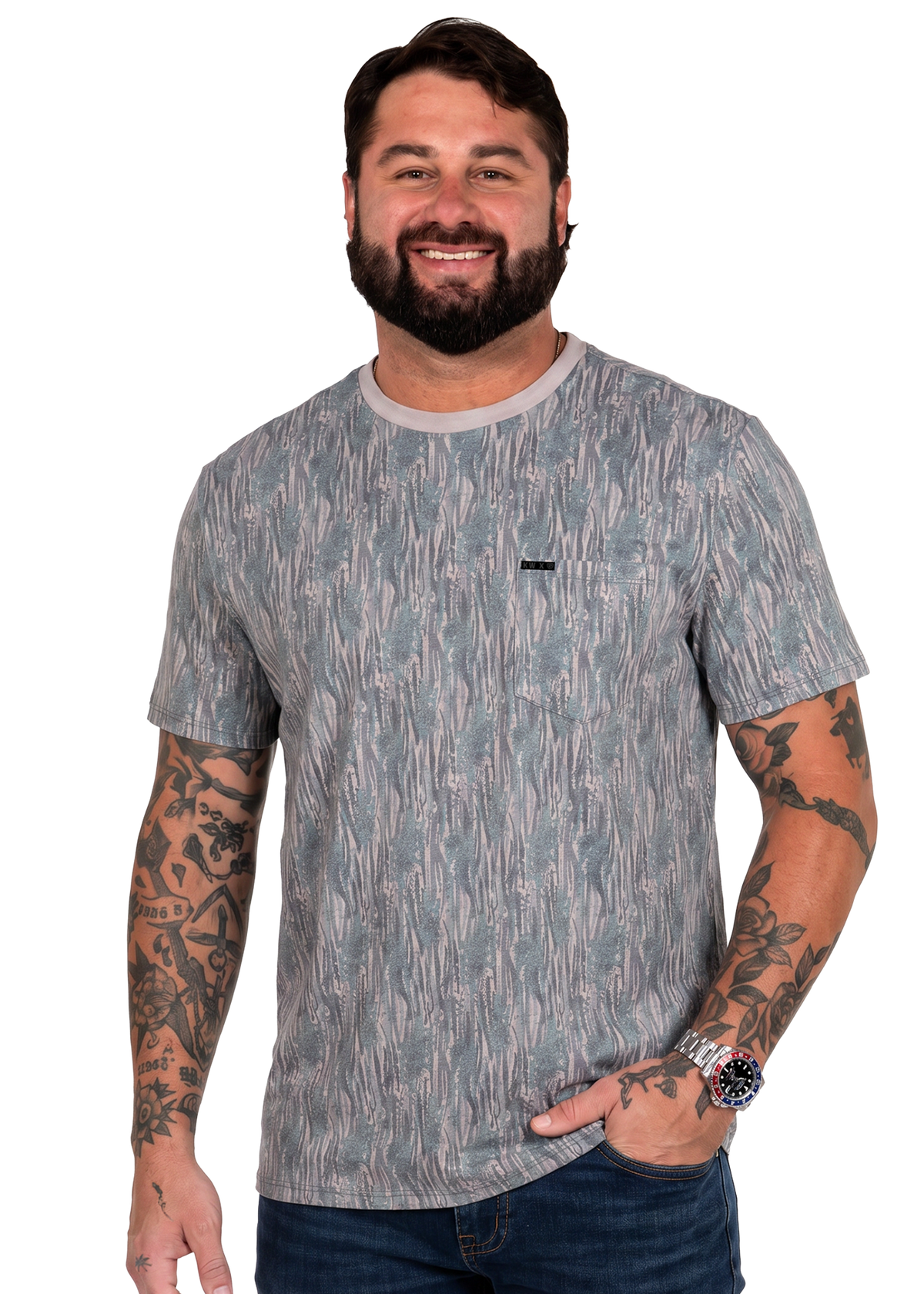 A smiling man with a beard and tattoos wearing a gray and blue abstract patterned short-sleeve t-shirt with a chest pocket, paired with dark blue jeans, against a plain white background.