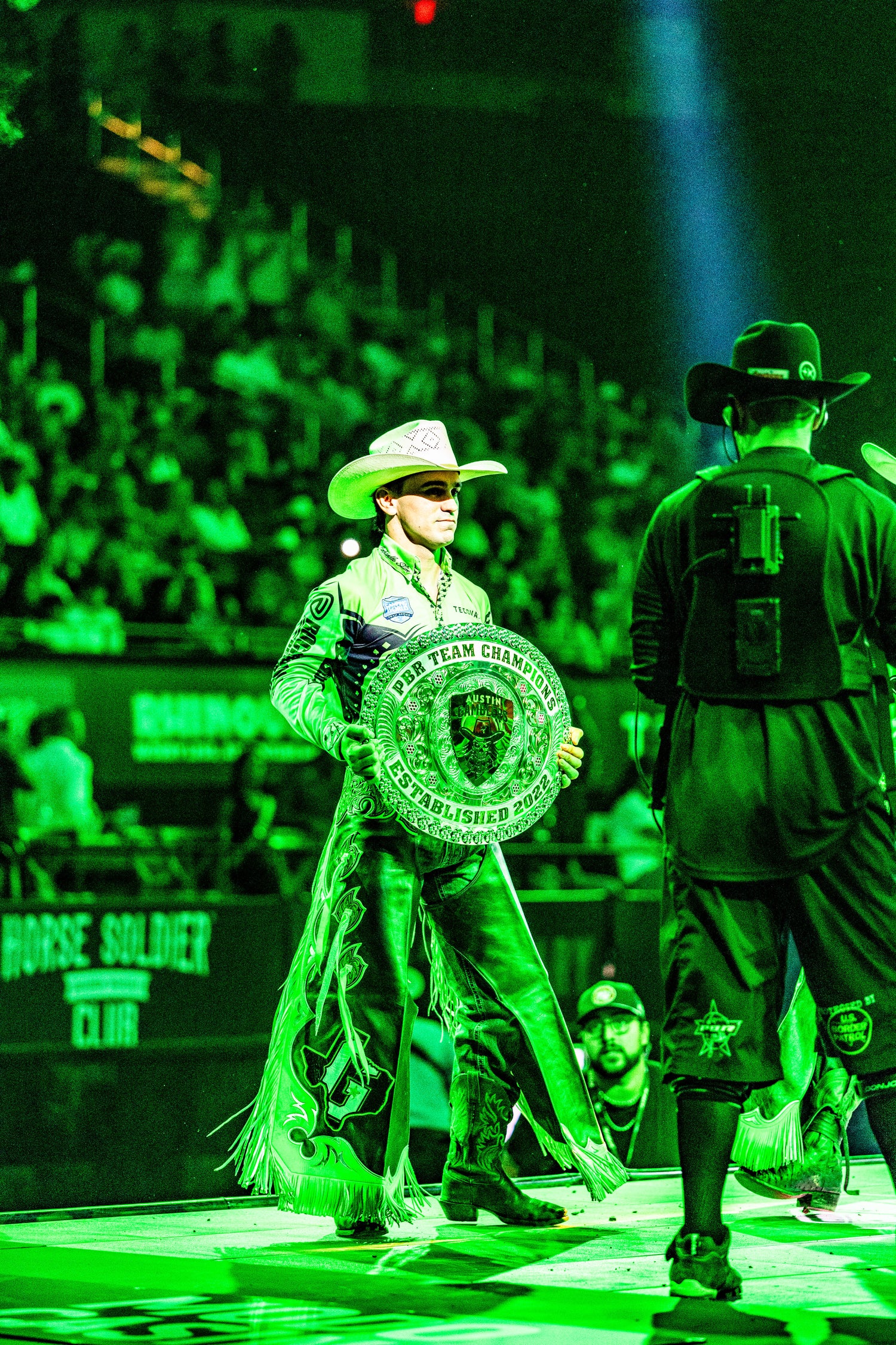 Person in cowboy attire holding a championship plaque on a stage with a crowd in the background.