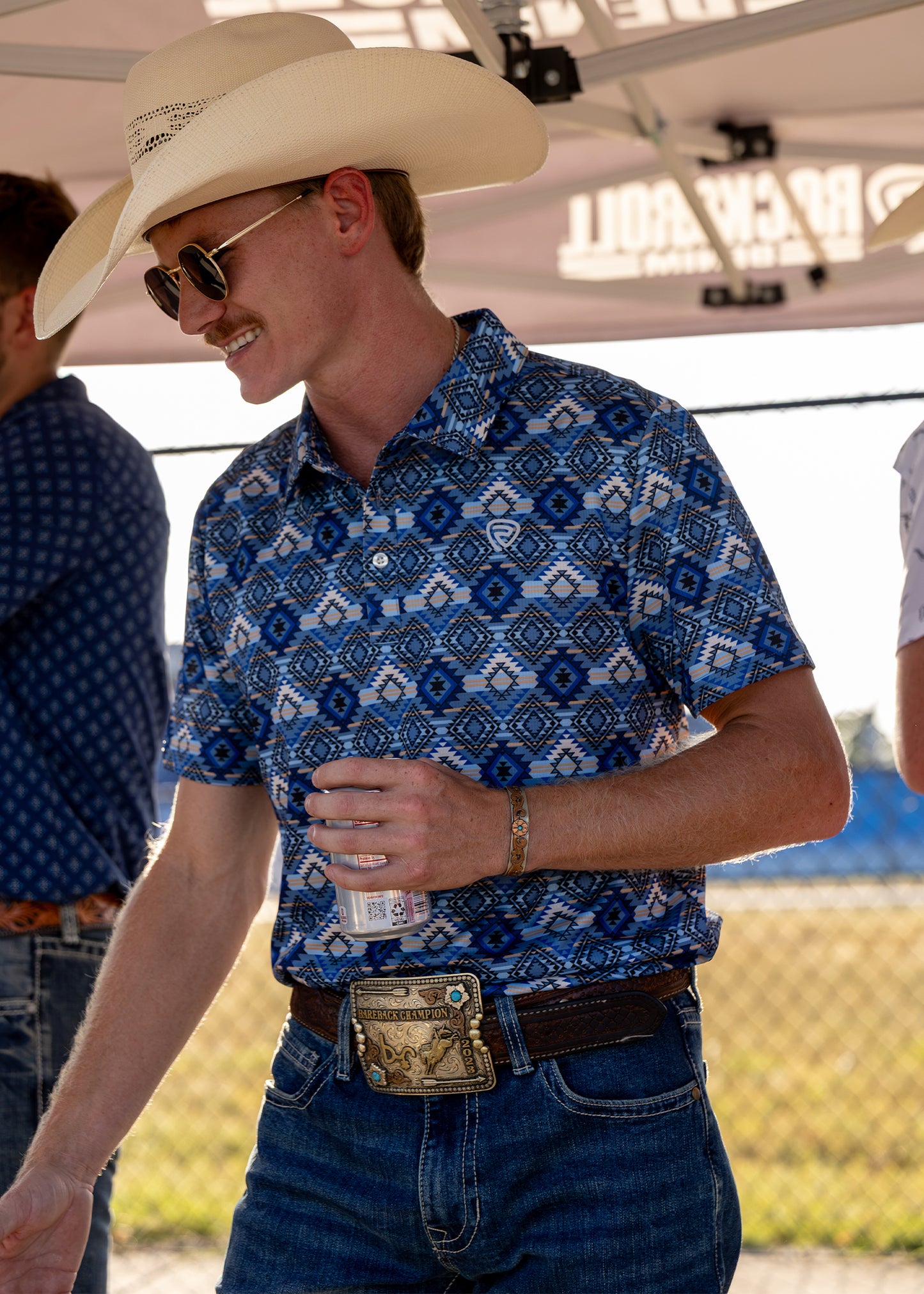 A man wearing a blue and white geometric patterned short-sleeve button-up shirt, blue jeans, standing under a white canopy tent.