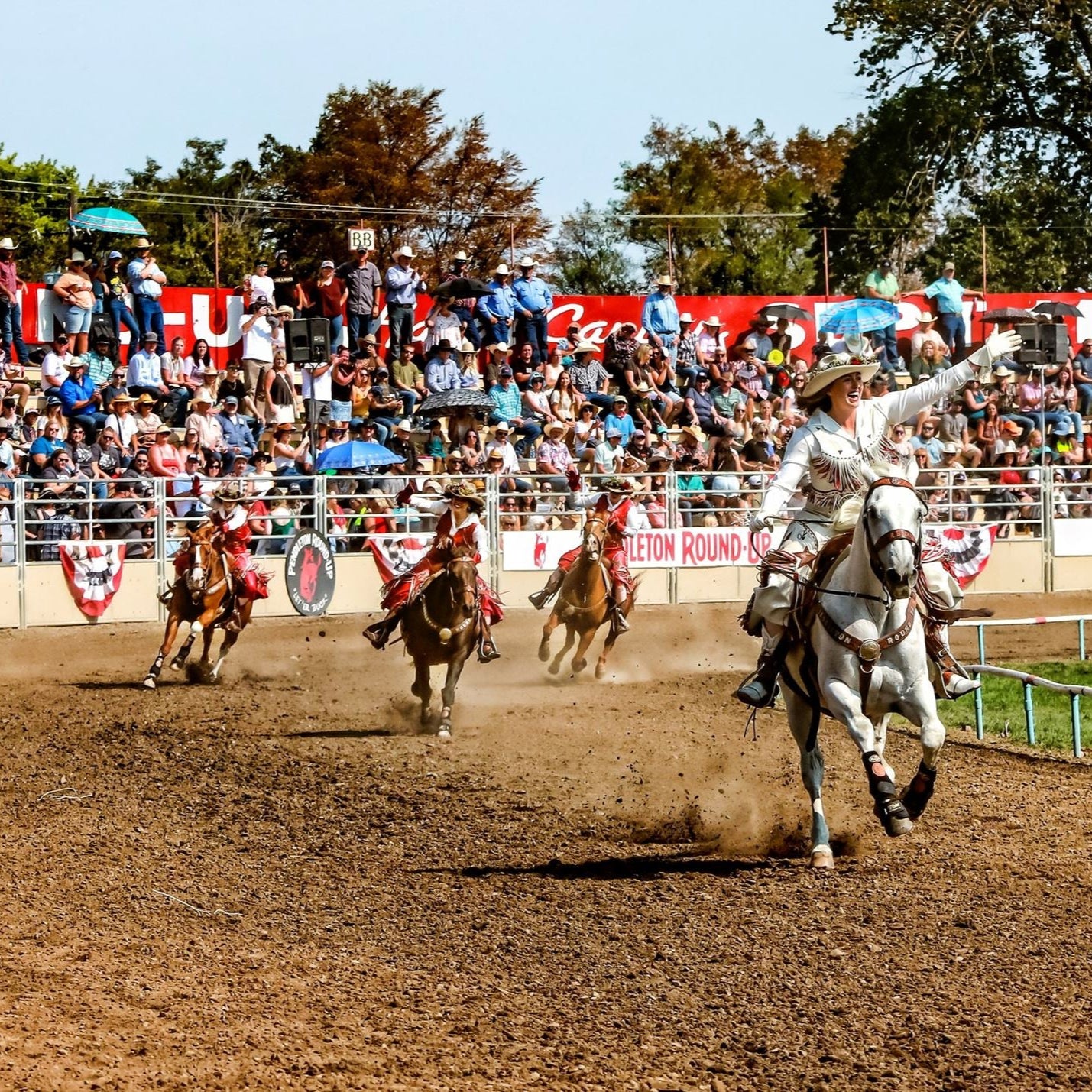 Rodeo riders perform in a dusty arena as a large crowd watches from the stands; one rider on a white horse raises an arm in the foreground.