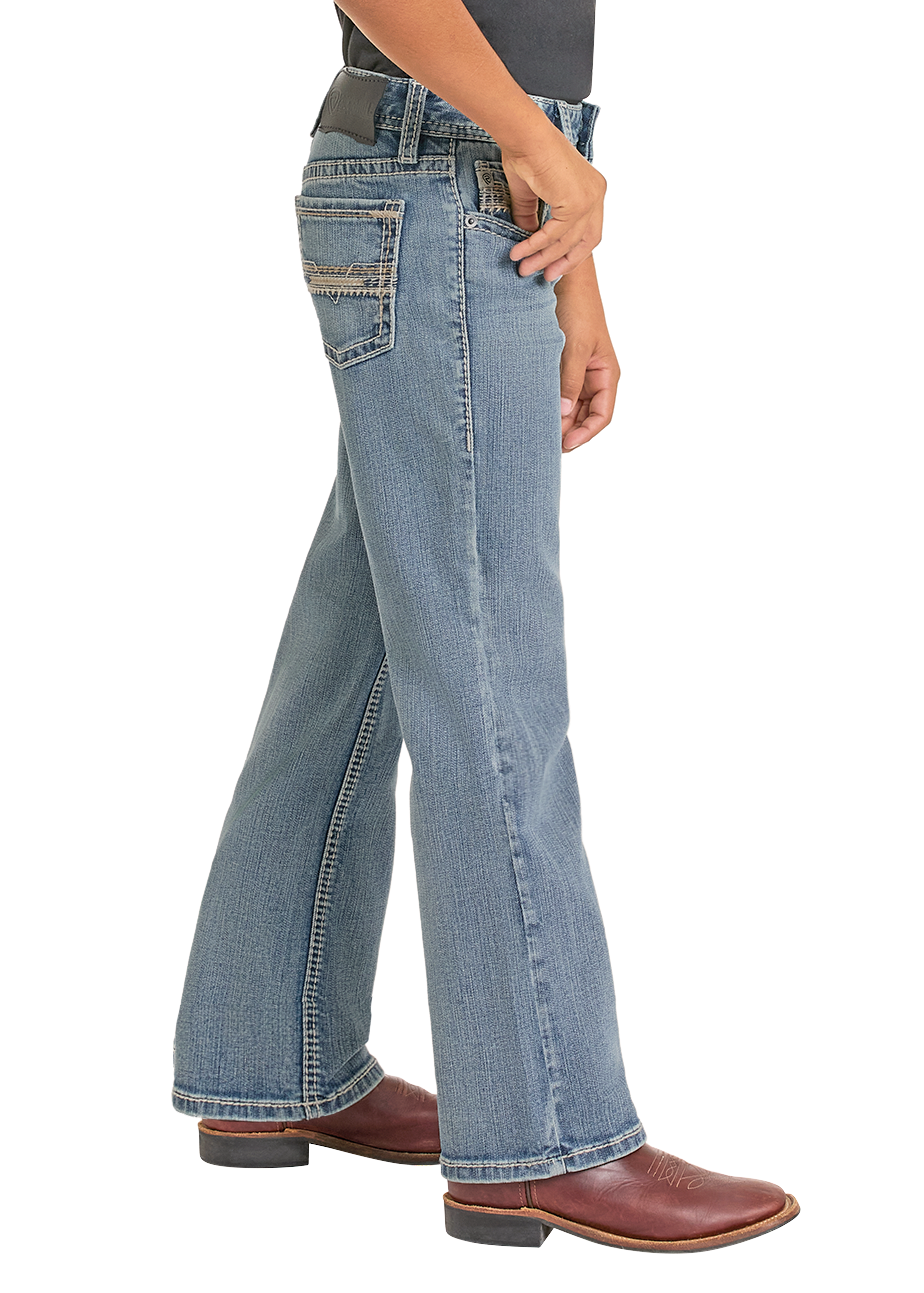 A person stands sideways with one hand in their pocket, wearing Rock & Roll Denim Boys' Regular Fit Light Feathered Stitch Bootcut Jeans and brown cowboy boots. The jeans feature decorative stitching on the back pocket.