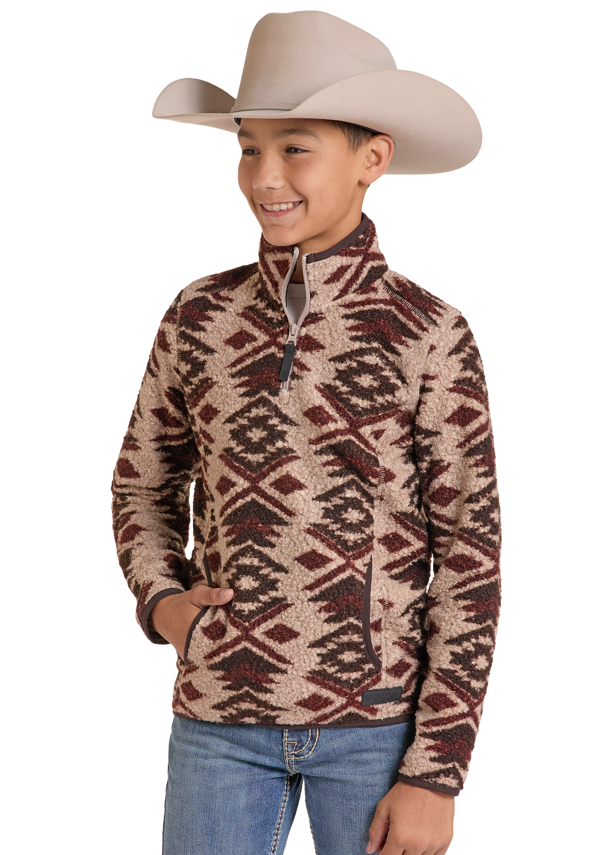 A young boy wearing a beige cowboy hat and a brown and beige geometric patterned quarter-zip fleece pullover sweater, paired with blue jeans, set against a plain white background.