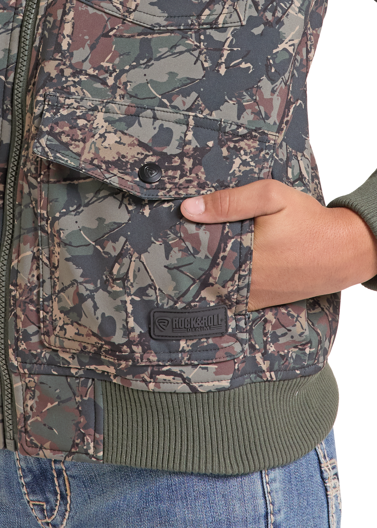 Close-up of a camouflage jacket with a front flap pocket, a hand in the pocket, and a visible Rocknroll brand patch on the lower front. The jacket has an olive green ribbed hem and cuffs, paired with blue jeans against a white background.