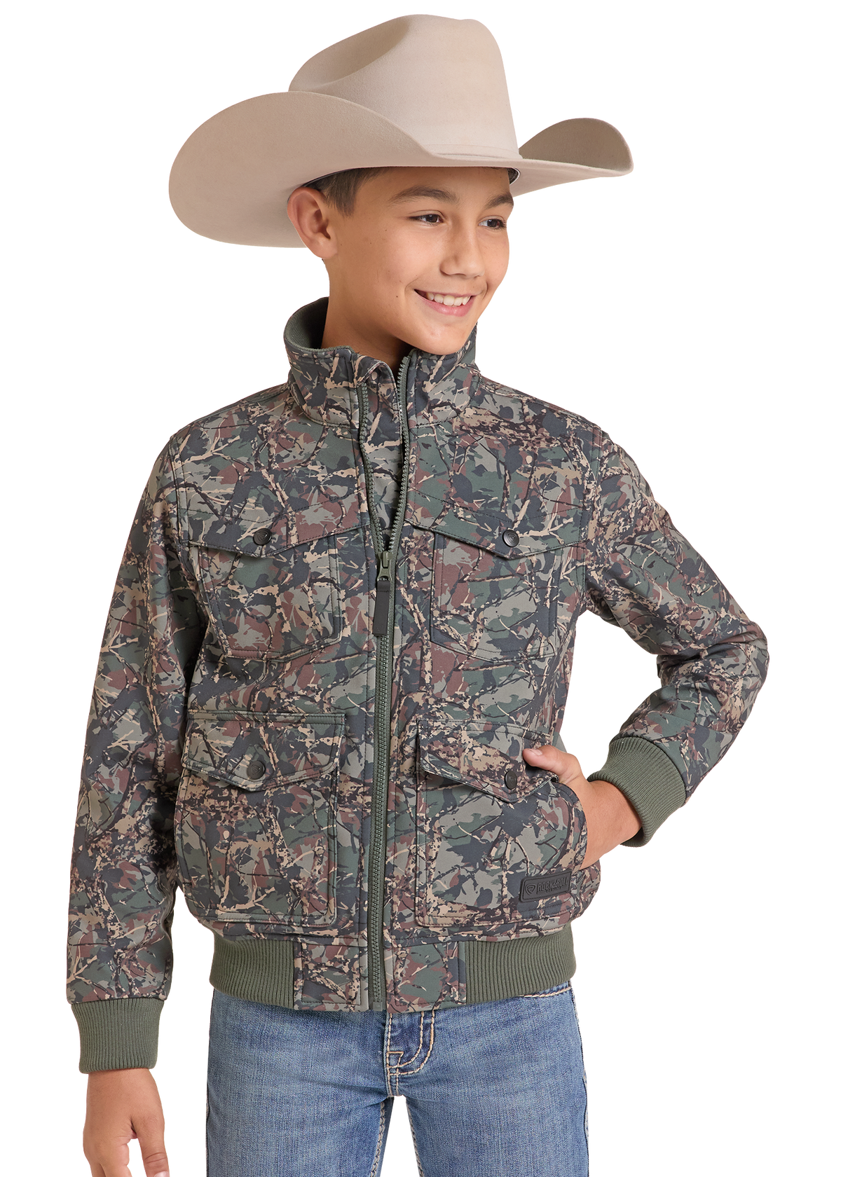 A young boy wearing a beige cowboy hat, a camouflage print zip-up jacket with green ribbed cuffs and hem, and blue jeans, photographed against a solid white background.