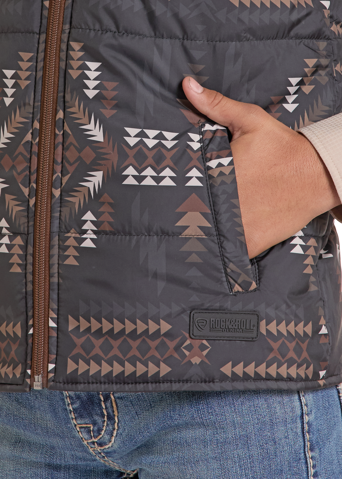 Close-up of a person wearing a dark gray puffer vest with a brown, beige, and white geometric pattern. A hand is tucked into a side pocket, and a Rockroll brand patch is visible near the bottom.