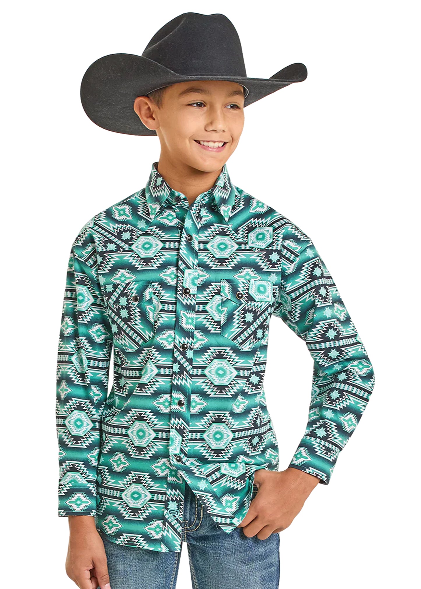 A smiling boy in a black cowboy hat, jeans, and a Rock & Roll Denim Boys' Jade Aztec Horizontal Print Long Sleeve Snap Shirt stands with one hand in his pocket, looking to the side.
