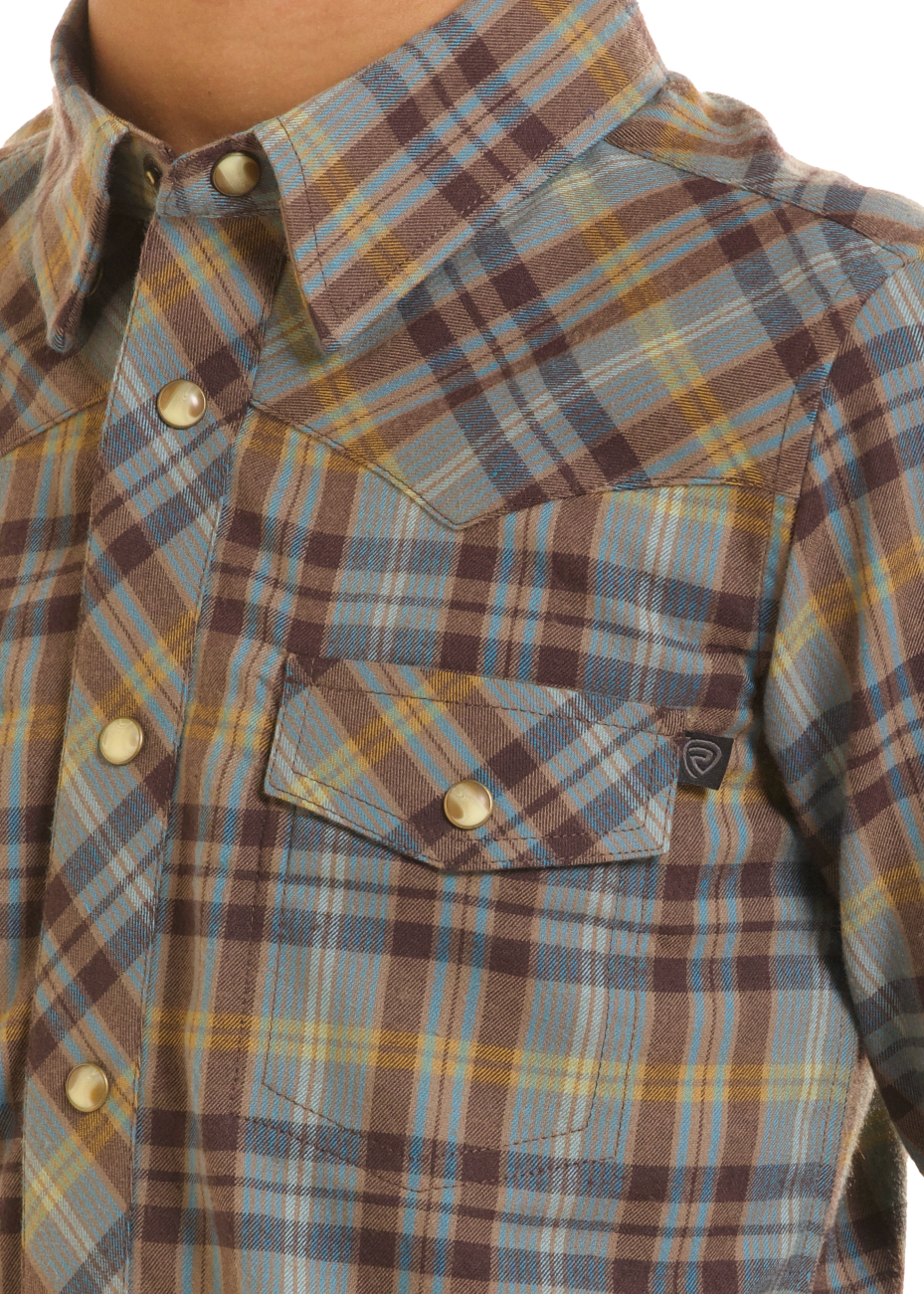 A close-up of a man wearing a long-sleeve plaid button-up shirt. The shirt features a pattern of muted blue, brown, yellow, and beige stripes, with a chest pocket and snap buttons.