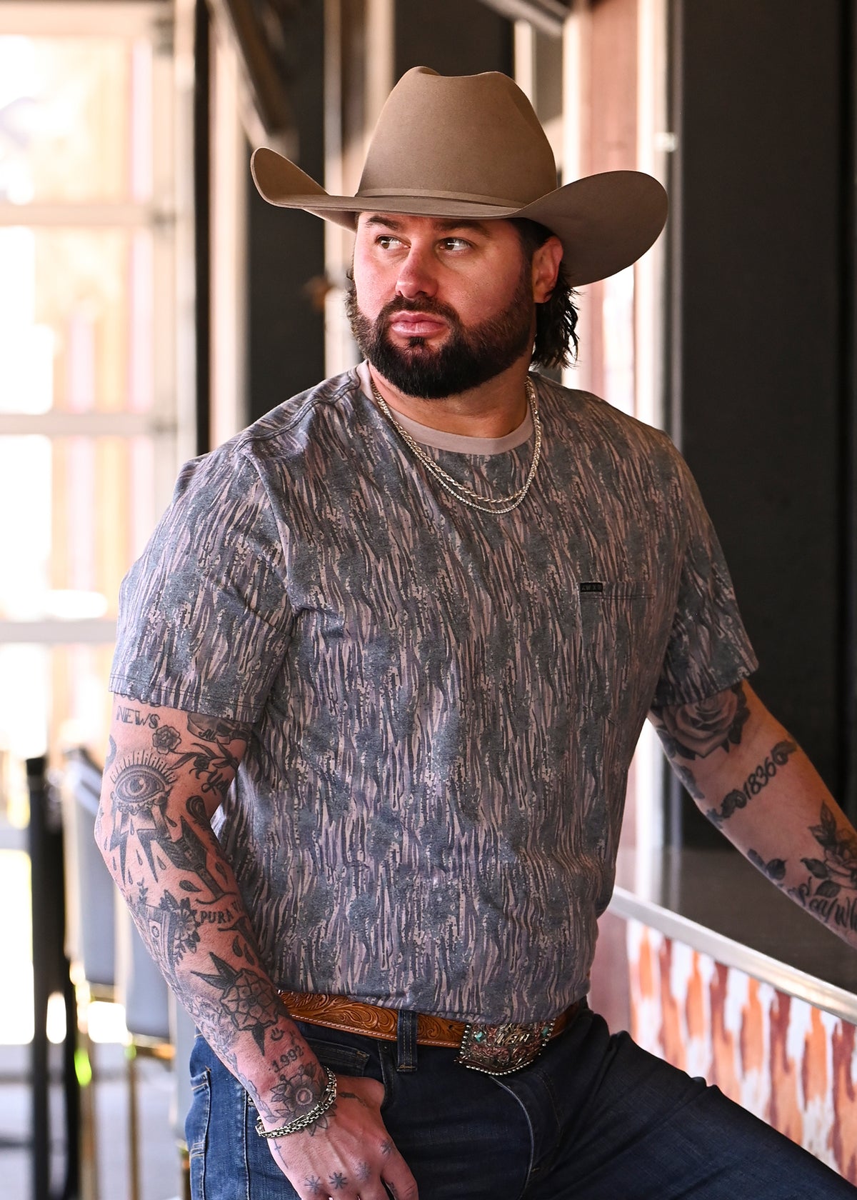 Koe Wetzel wearing a cowboy hat, a camo short-sleeve t-shirt, and jeans, leaning against a bar counter. 