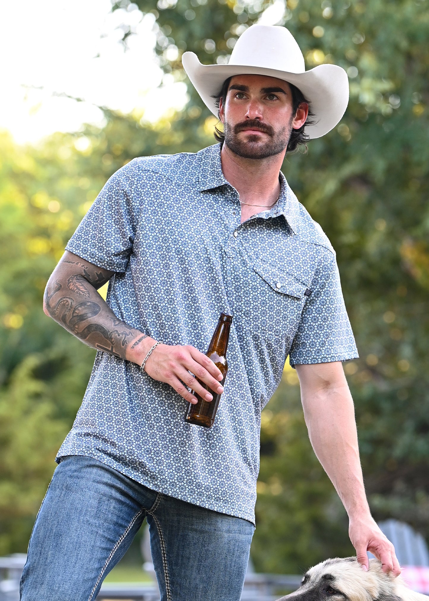 A man wearing a white cowboy hat, a blue and blue patterned polo, and blue jeans, holding a brown beer bottle and touching a dog outdoors.