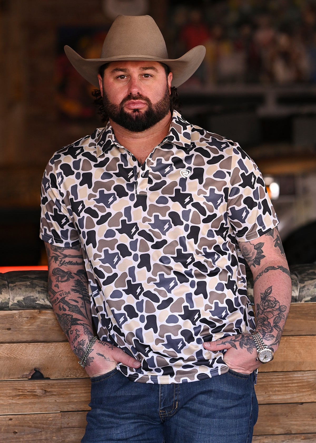Koe Wetzel wearing a Texas print camo polo with jeans and a cowboy hat in a bar setting. 