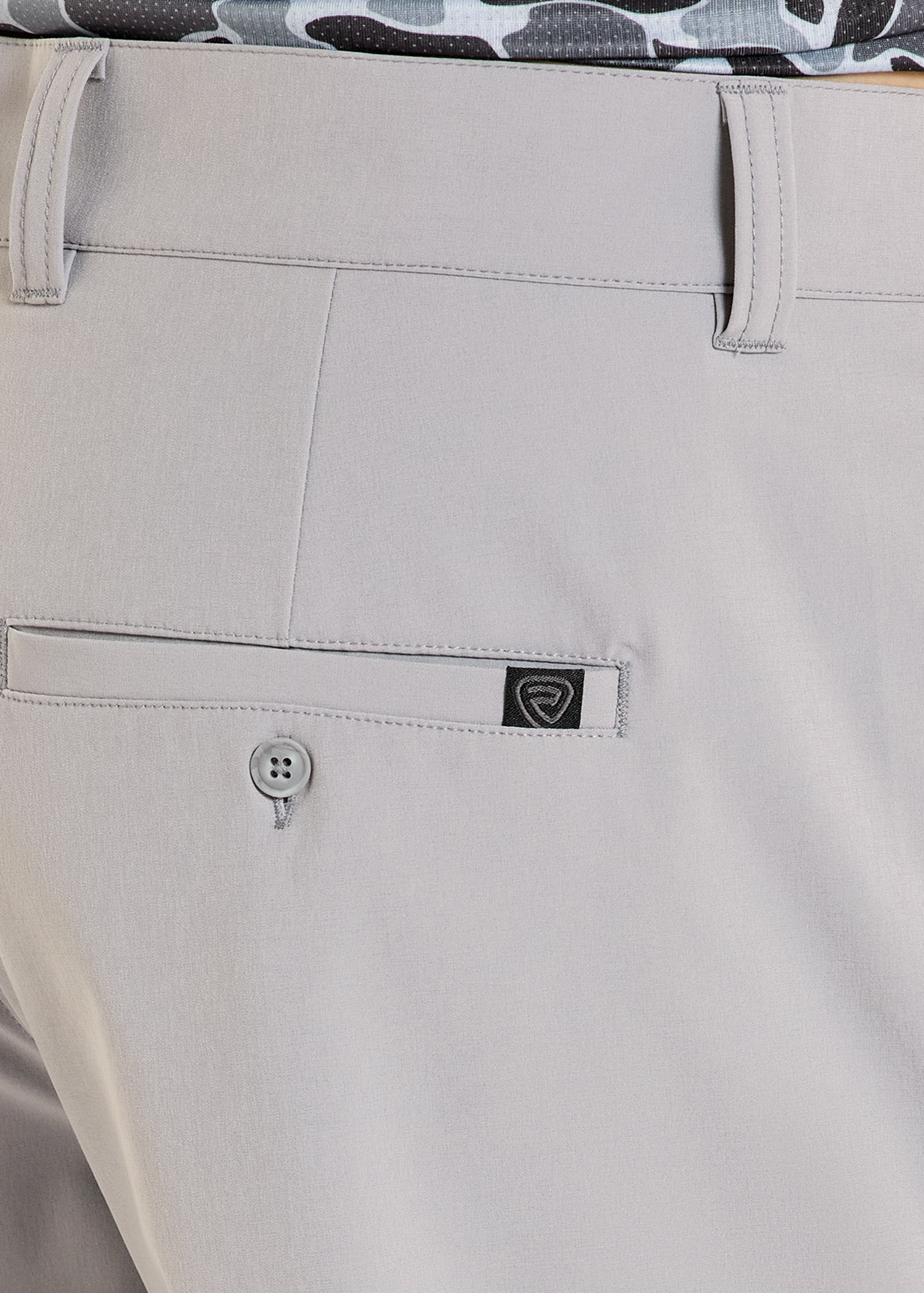 Close-up of beige pants with a brand logo on the pocket, featuring a camouflage shirt in the background.