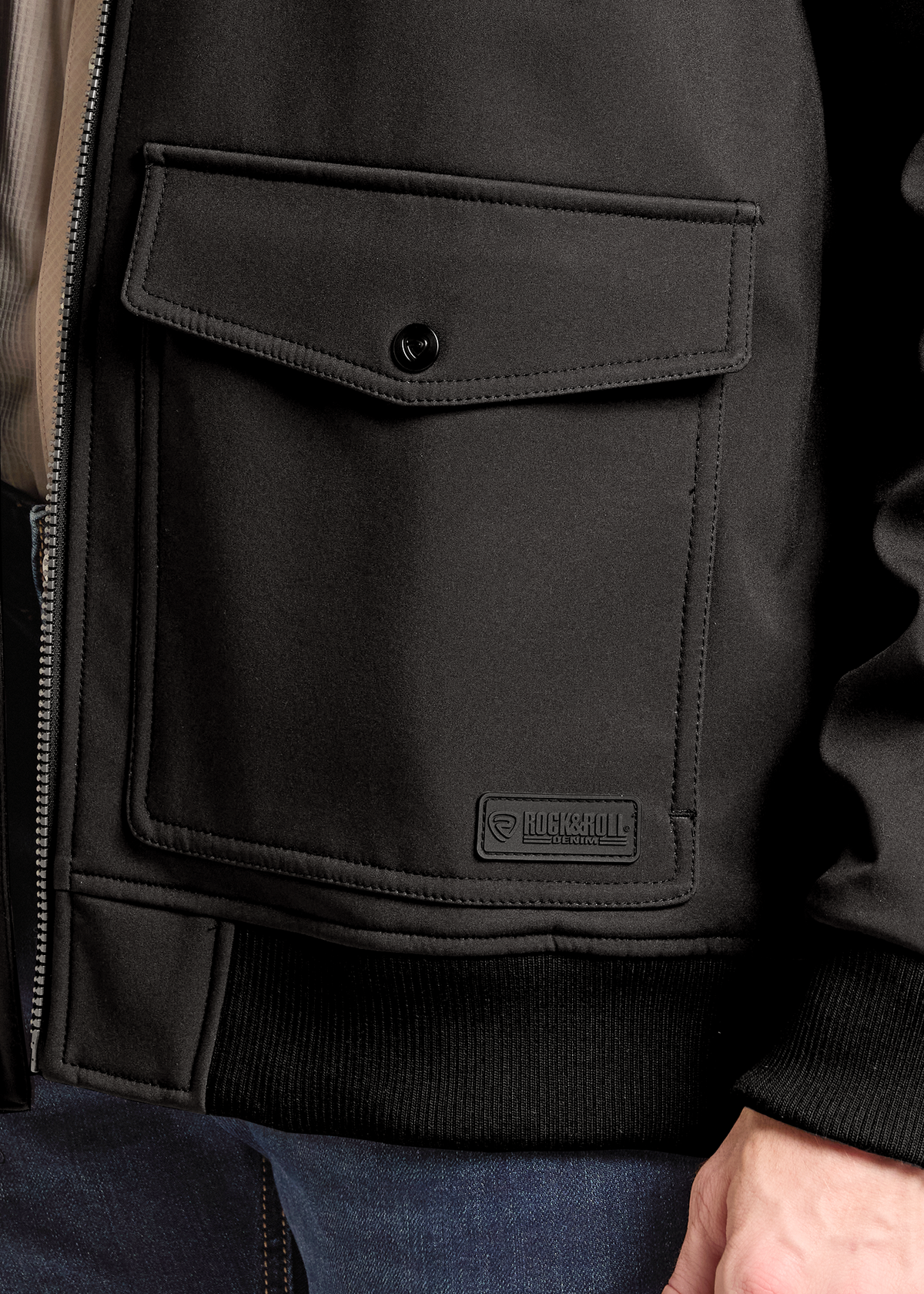 Close-up of a black jacket chest pocket with a snap tab and a small rectangular logo patch reading Barbour, worn by a person in blue jeans.