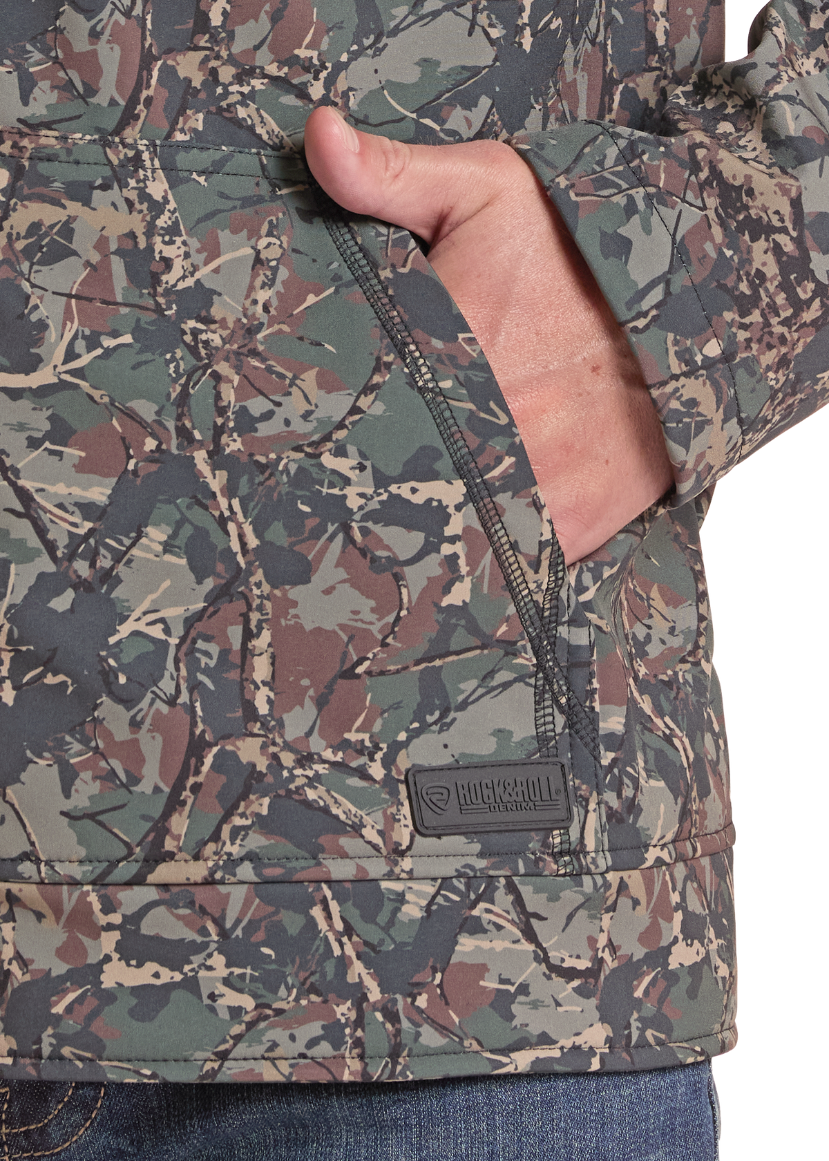Close-up of a camouflage jacket with a hand in a front zip pocket, showing a small black Buckshot brand patch near the hem.