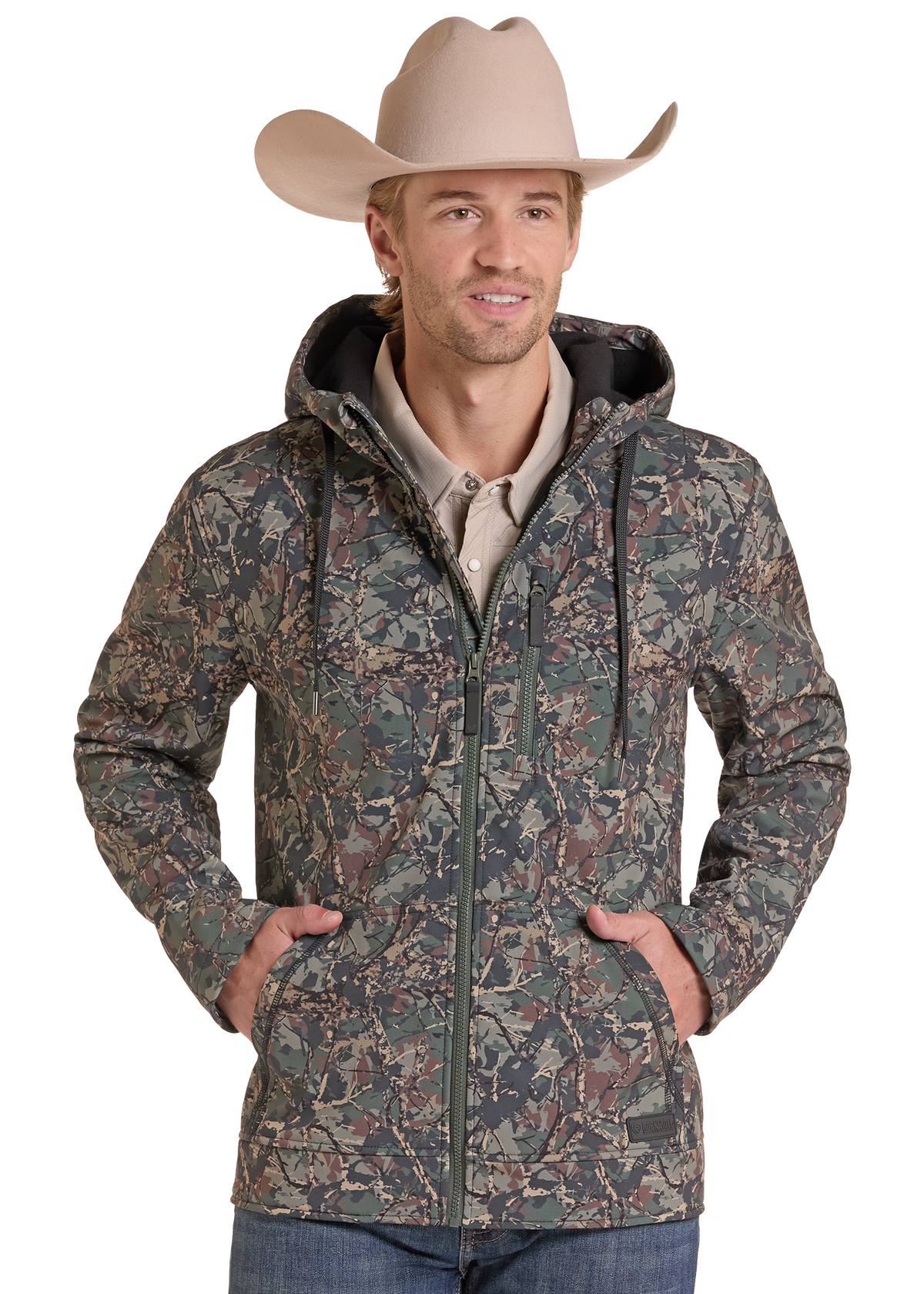 A man wearing a camouflage hooded jacket, a beige cowboy hat, and a light beige shirt, standing against a white background with his hands in the pockets of the jacket.