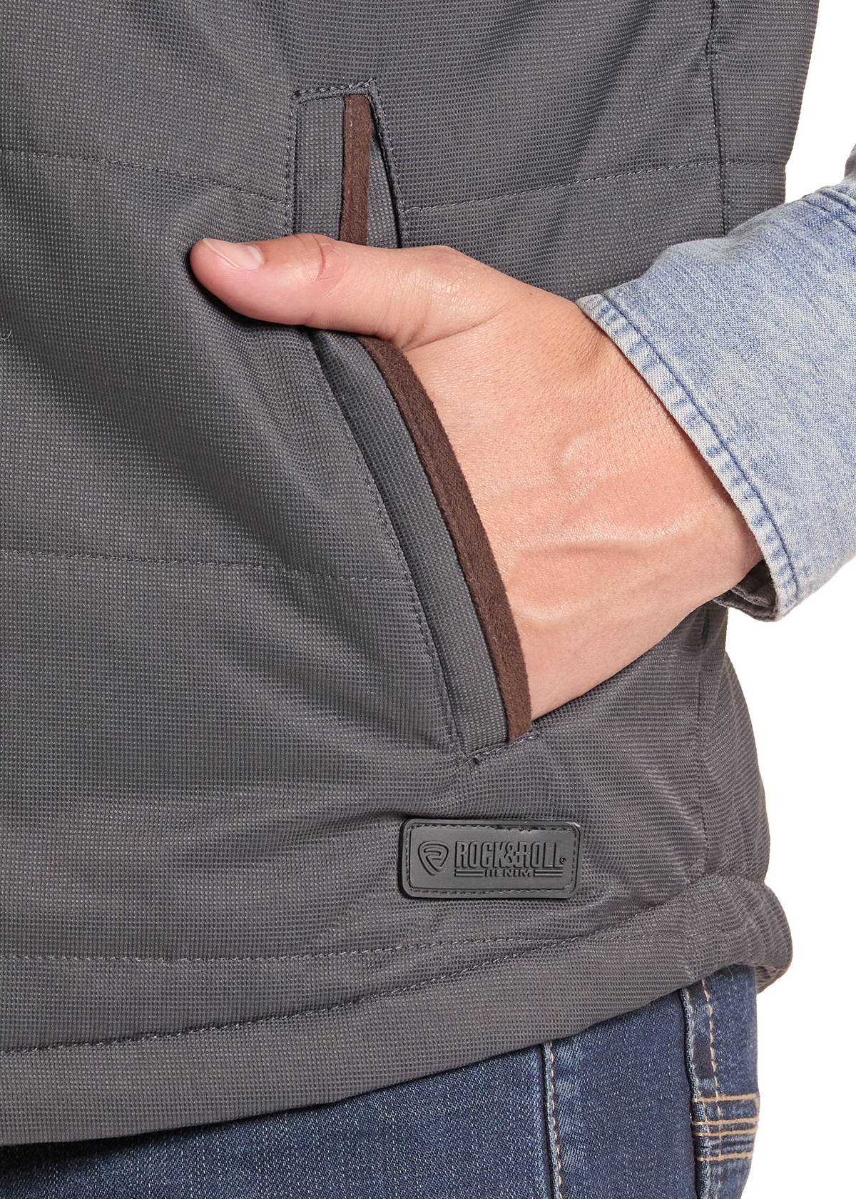 Close-up of a person wearing a dark gray quilted vest with a side pocket, hand in pocket, and a Bucknell logo patch on the lower front. A light blue denim shirt cuff is visible.