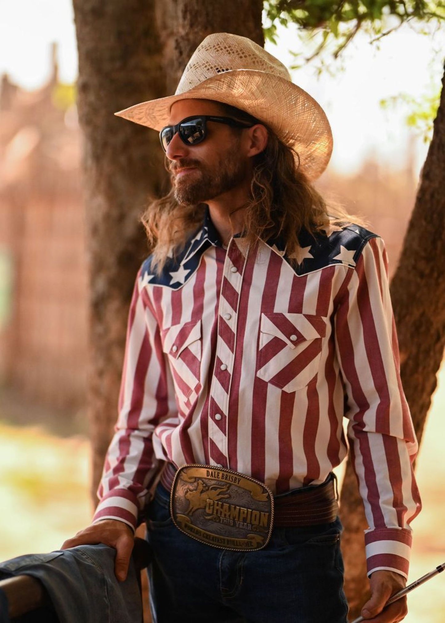 Dale Brisby wearing a cowboy hat, sunglasses, and a patterned shirt with a 'Champion' belt buckle.