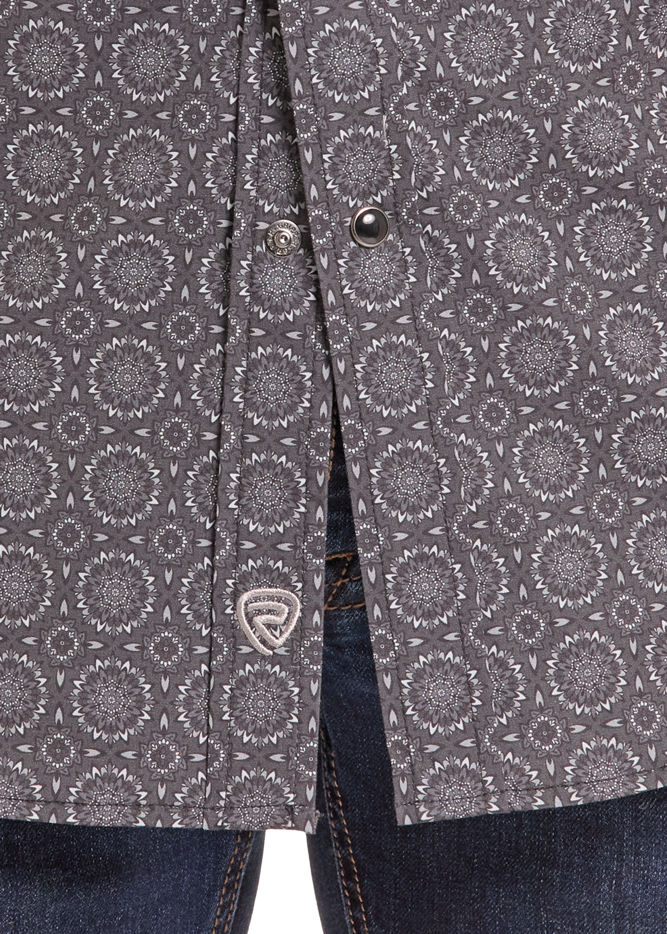Close-up of a dark gray patterned button-up shirt with a circular medallion print, styled with blue jeans. A silver shield logo is visible on the shirt's button placket.