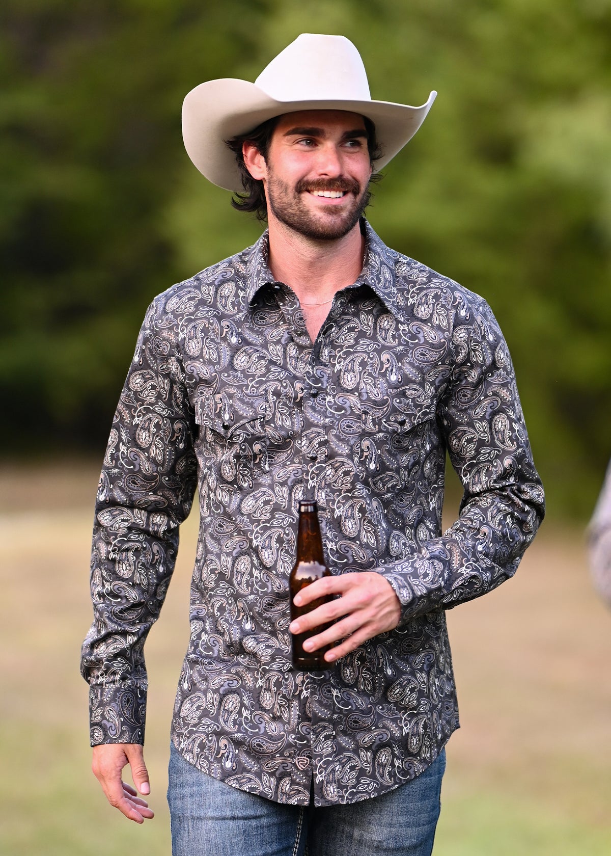 A man wearing a white cowboy hat and a long-sleeve snap shirt with a gray and black paisley pattern, holding a brown bottle outdoors.
